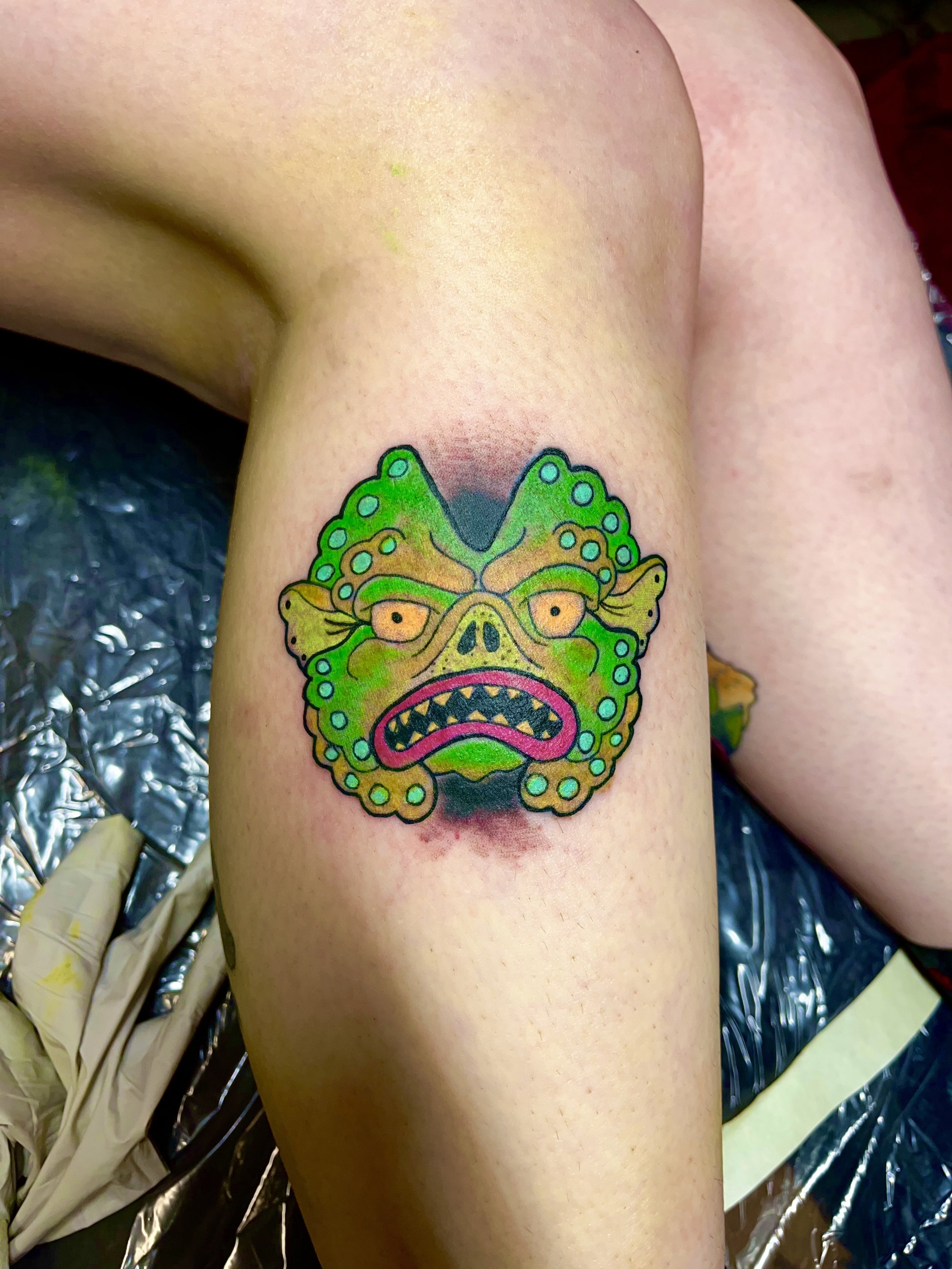 Colorful tattoo of a cartoonish creature with a face resembling a frog or goblin, featuring multiple eye-like spots, sharp teeth, and bright green and yellow coloring, on a person's thigh.