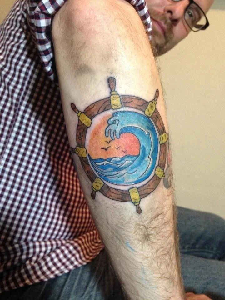 A tattoo of a beach scene with a sunset, ocean waves, and birds, surrounded by a wooden boat ring with yellow lights.
