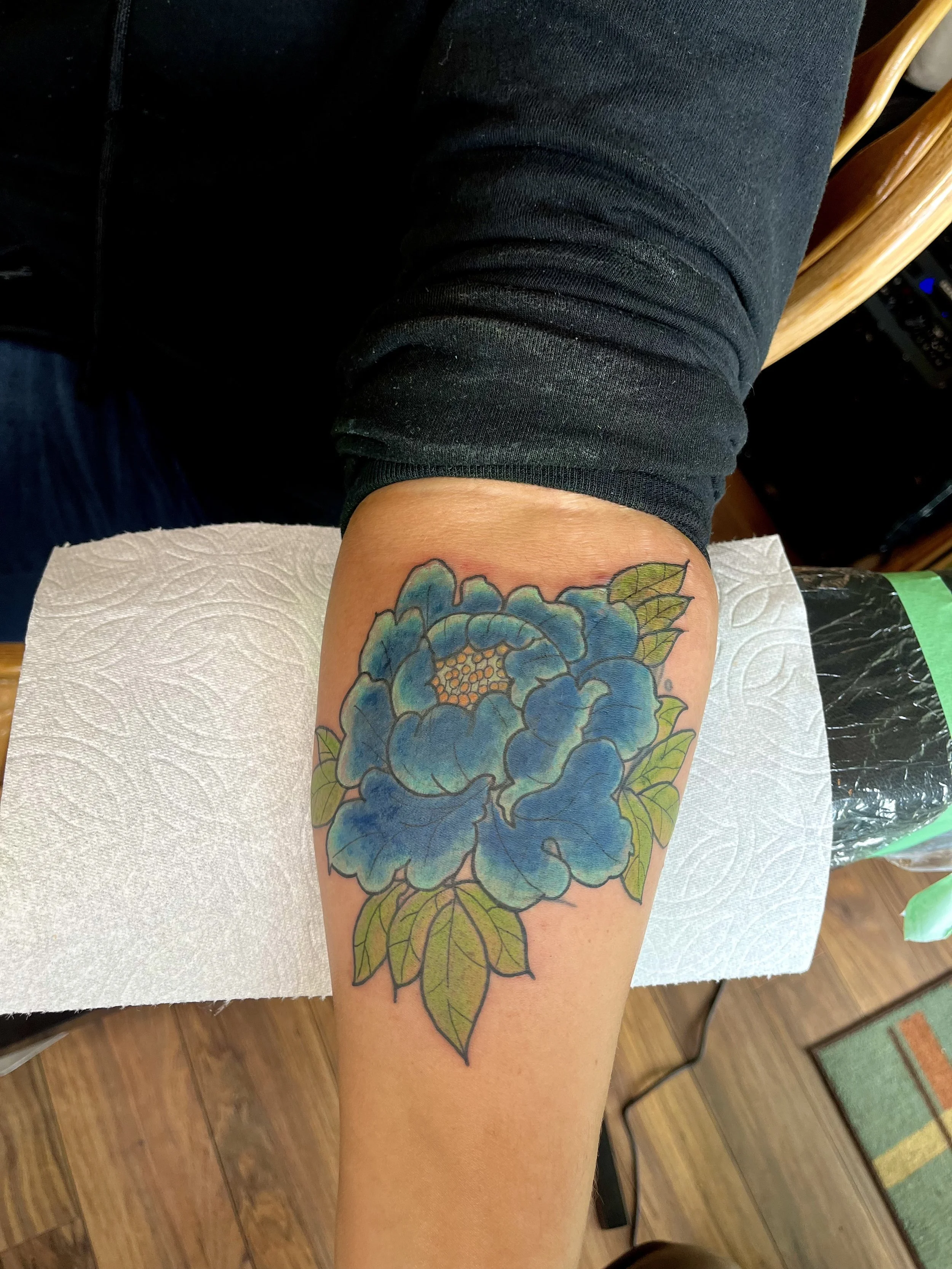 Colorful tattoo of a blue flower with green leaves on a person's forearm.