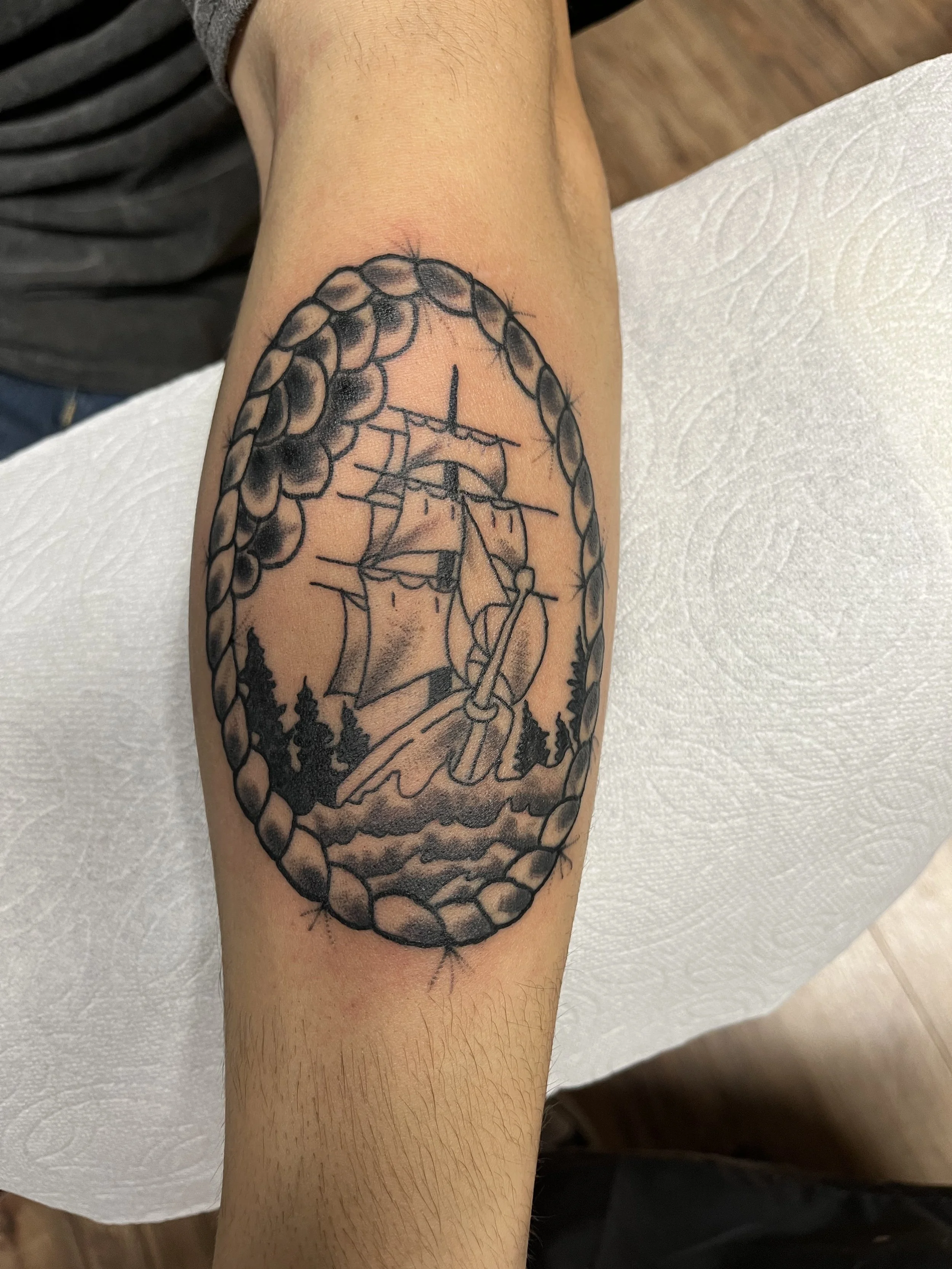 Tattoo of a sailing ship within an oval rope border, with trees and waves below, on a person's forearm.