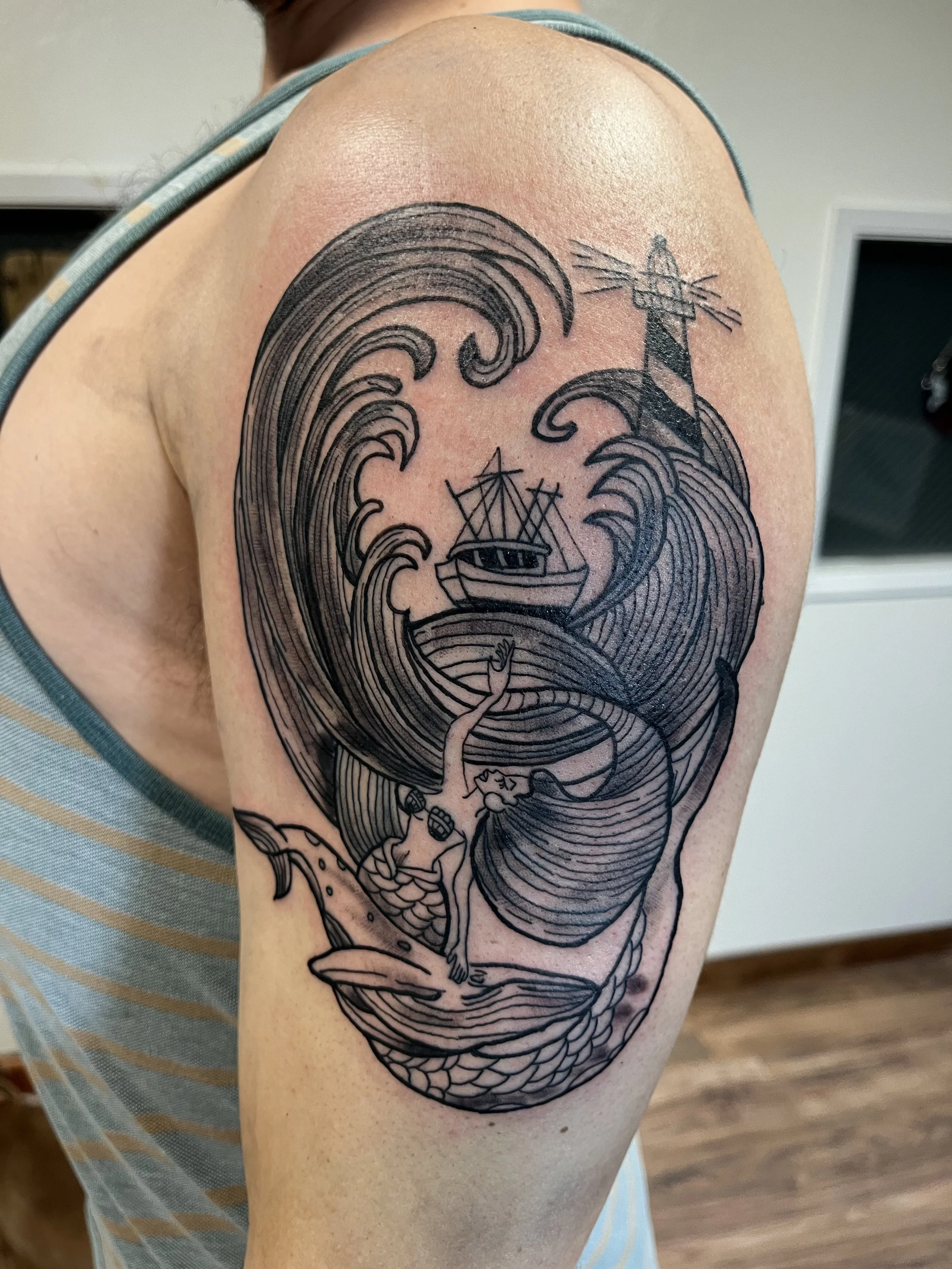 Black tattoo of a mermaid riding a fish, with large crashing waves, a boat in the waves, and a lighthouse in the background.