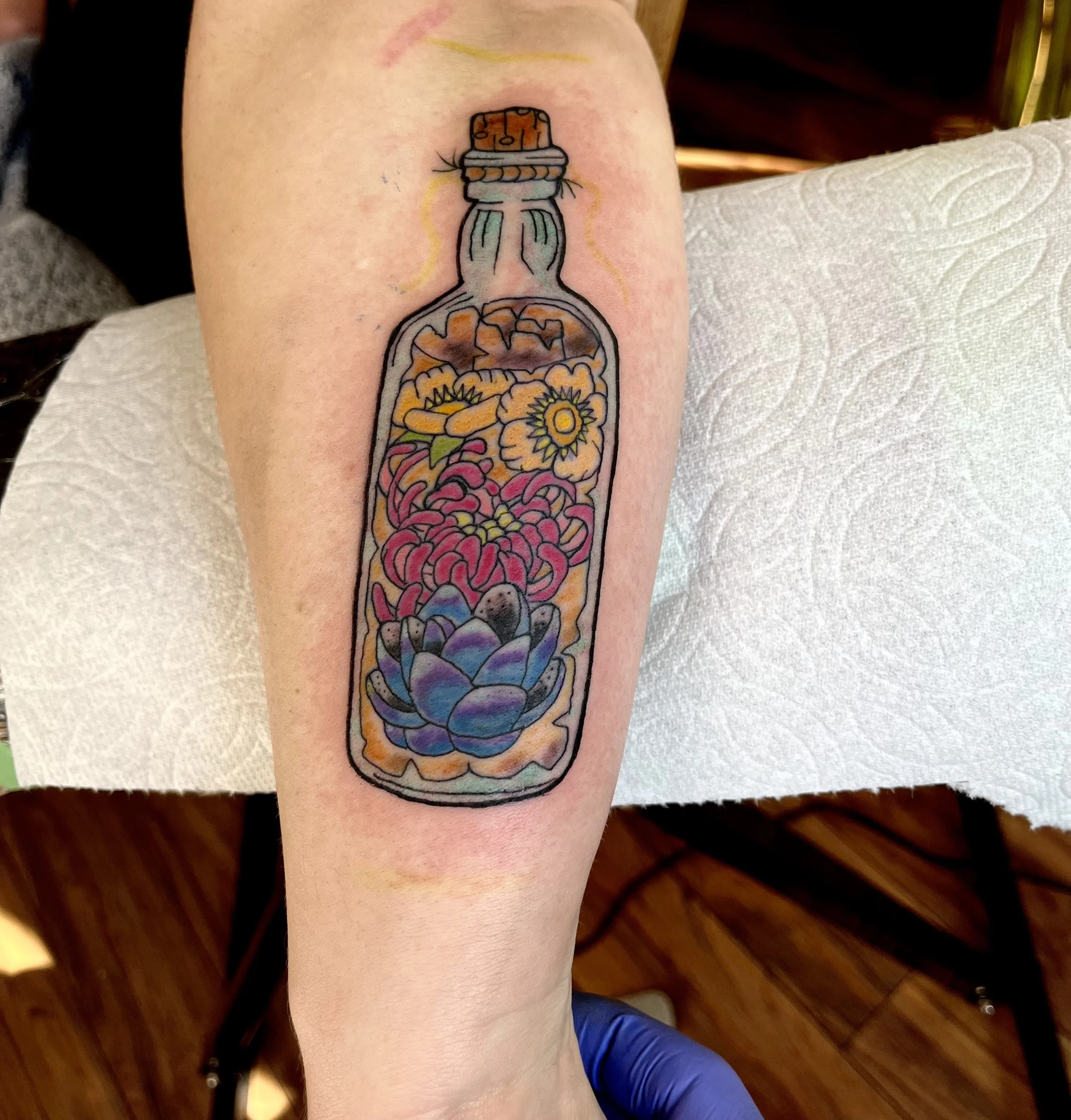 Colorful tattoo of a bottle filled with various flowers and a succulent, with the bottle sealed with a cork.