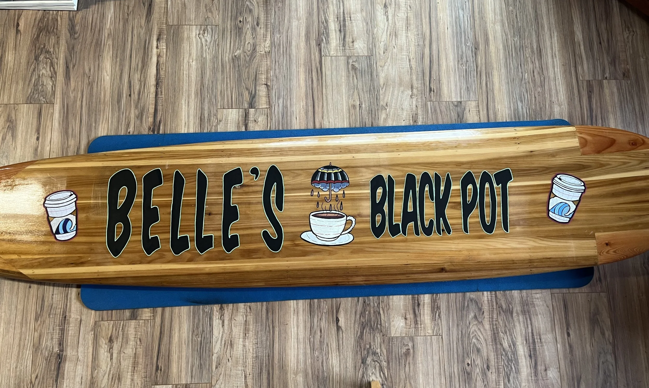 A wooden surfboard-shaped sign that reads "Belle's Black Pot" with illustrations of coffee cups and a coffee umbrella, placed on a wooden floor.