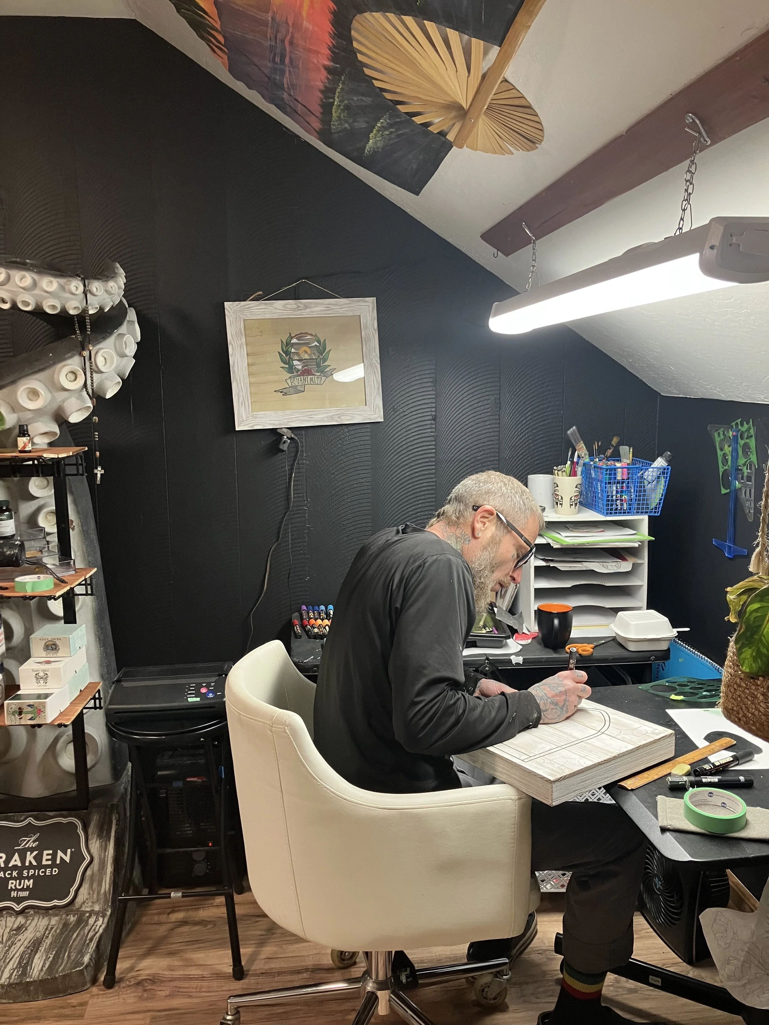 A man with tattoos and glasses sitting at a art desk, working on a wooden surface, in a cozy, decorated room.