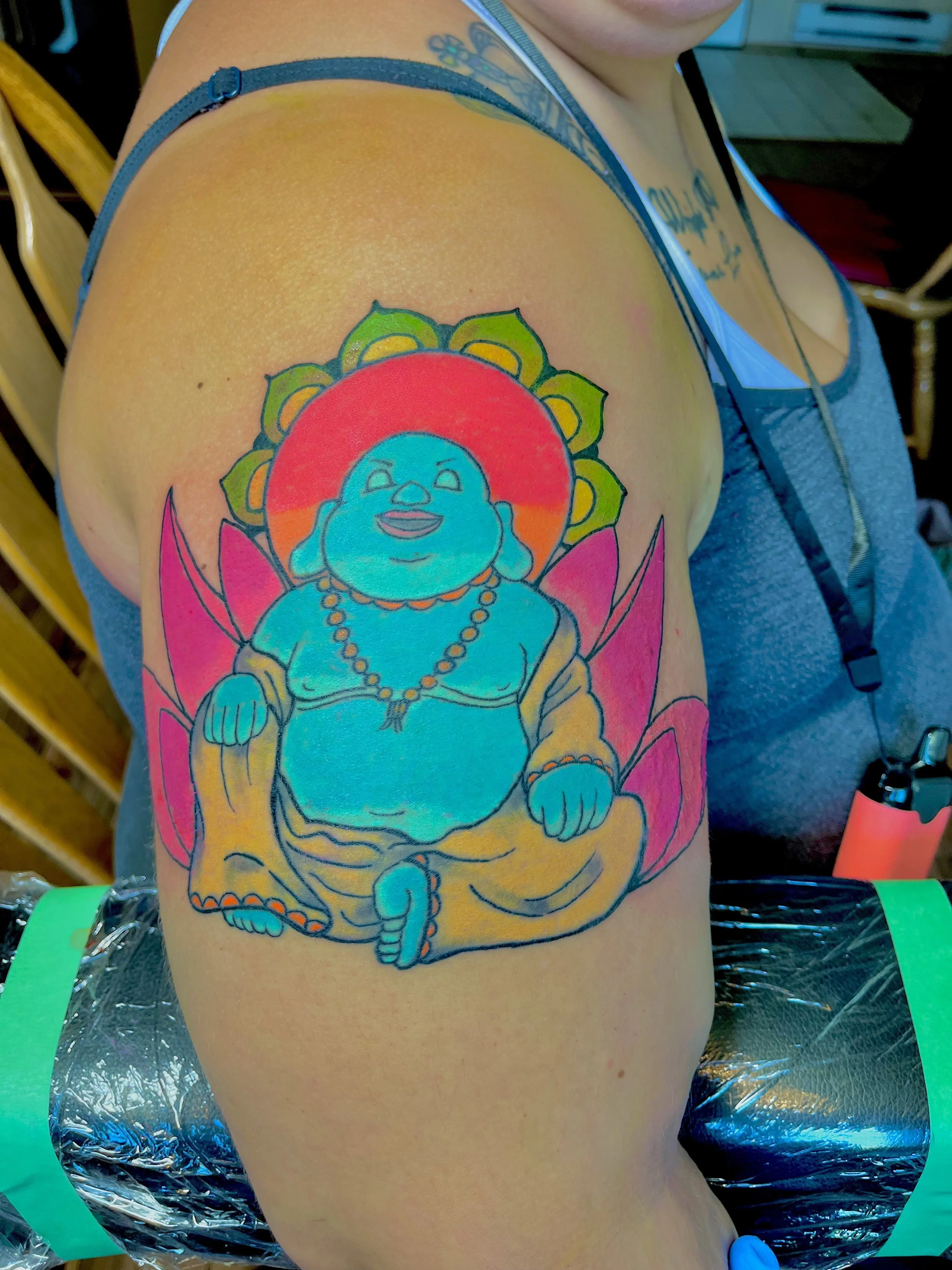 Colorful tattoo on person's upper arm depicting a cartoon-like, joyful, smiling figure with a round belly, sitting cross-legged with a beaded necklace, in front of a large red-orange sun and pink and green lotus petals.