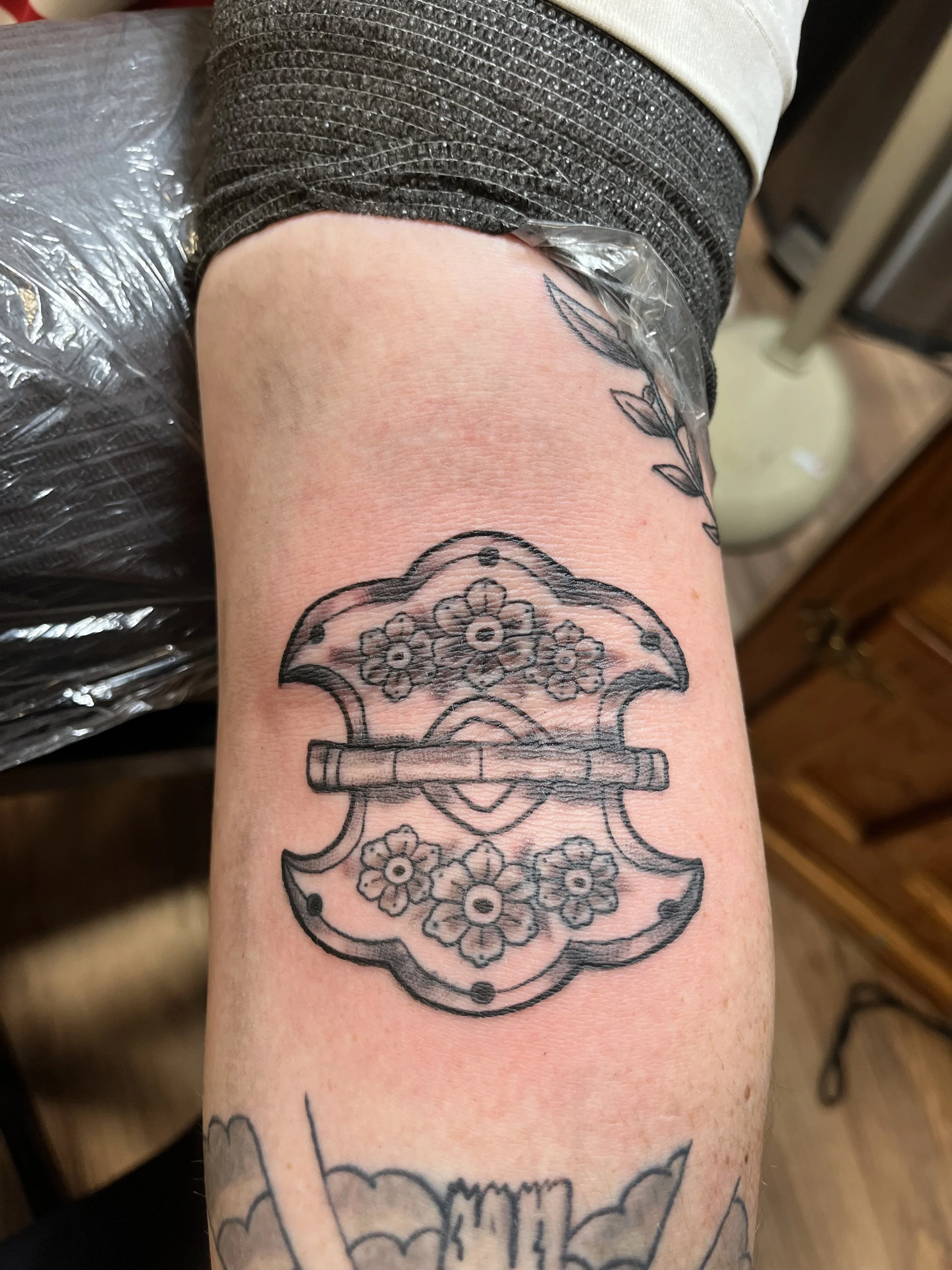 Outline of a floral and decorative tattoo design on a person's forearm, featuring a symmetrical floral pattern and a horizontal bar with a curved, ornate background.