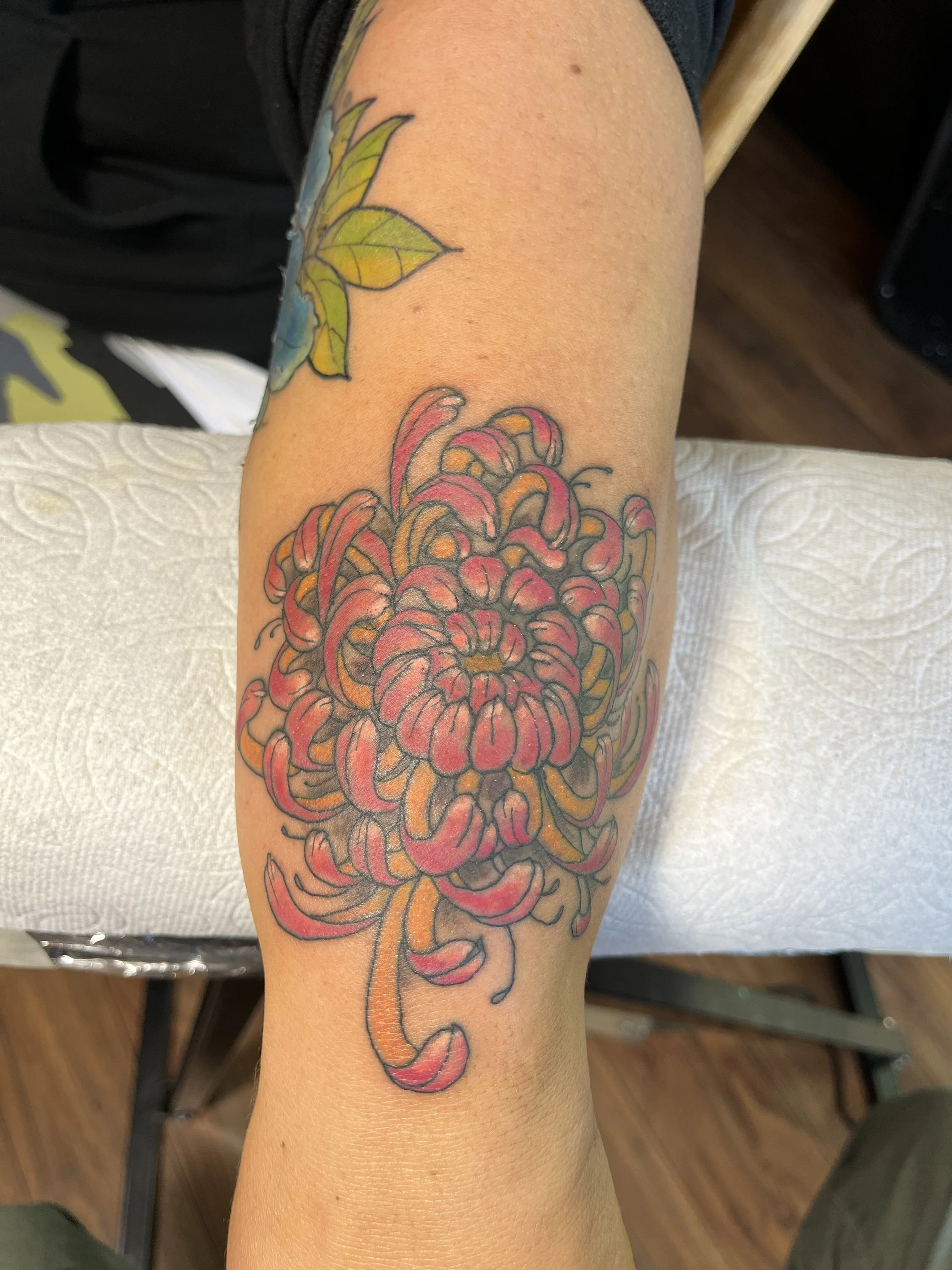 Colorful tattoo of a large flower on a person's forearm, with petals in shades of pink and orange, and a smaller flower tattoo above it with green and yellow leaves.