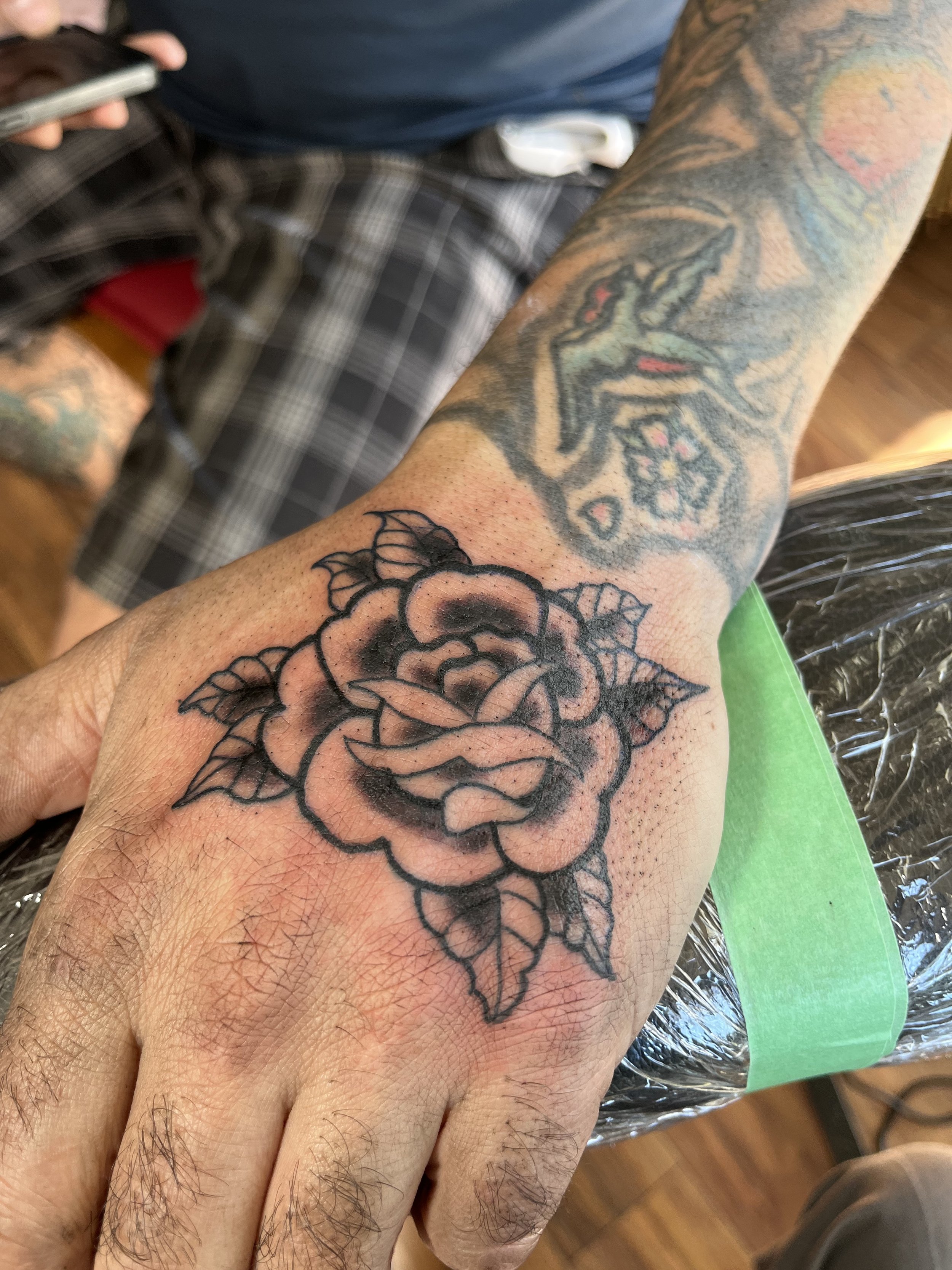 Black outline tattoo of a rose on a person's hand with green leaves and shading, on the person's finger.