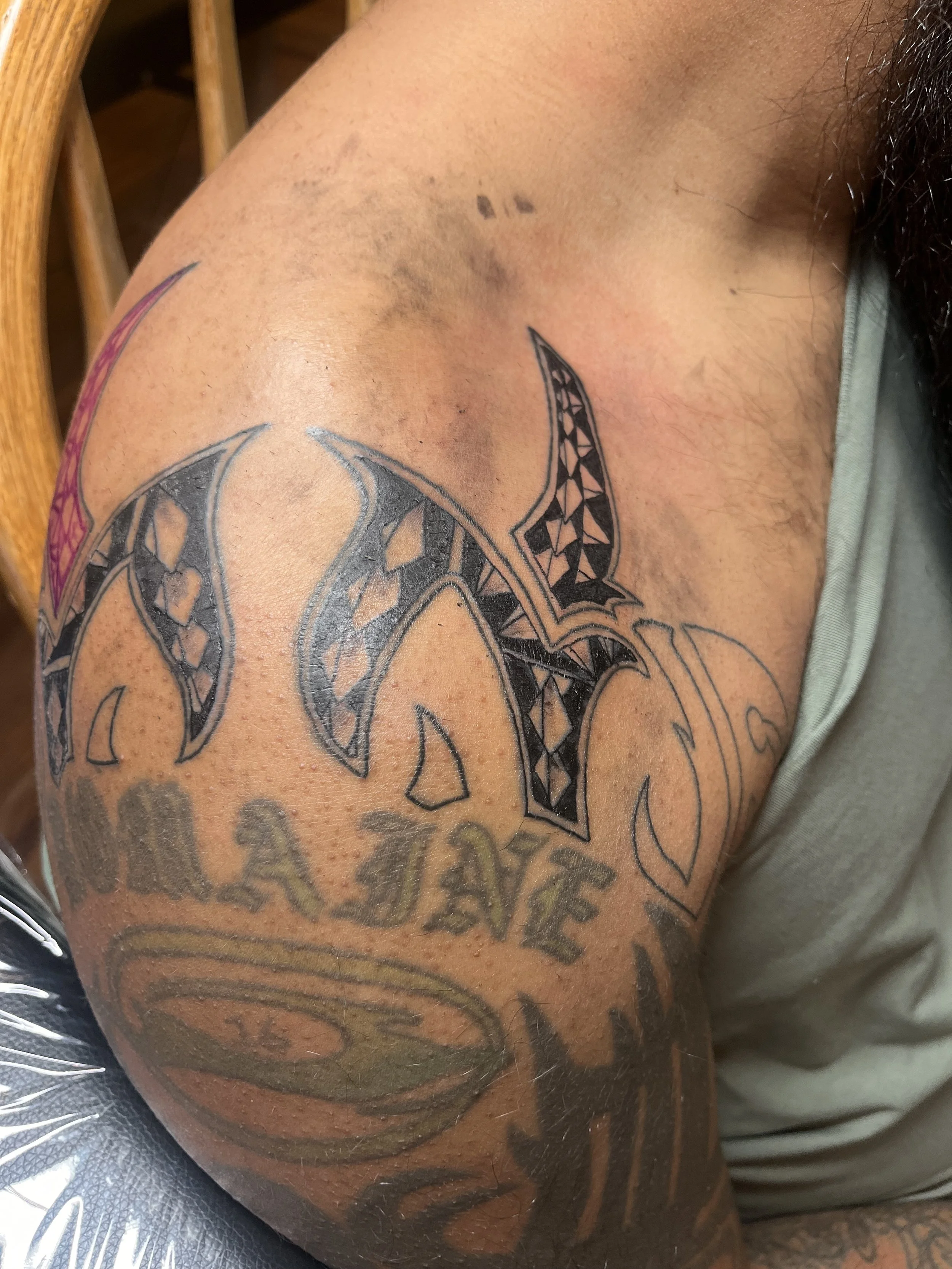 Close-up view of a person's upper arm and shoulder with a tattoo featuring stylized black tribal patterns and text.
