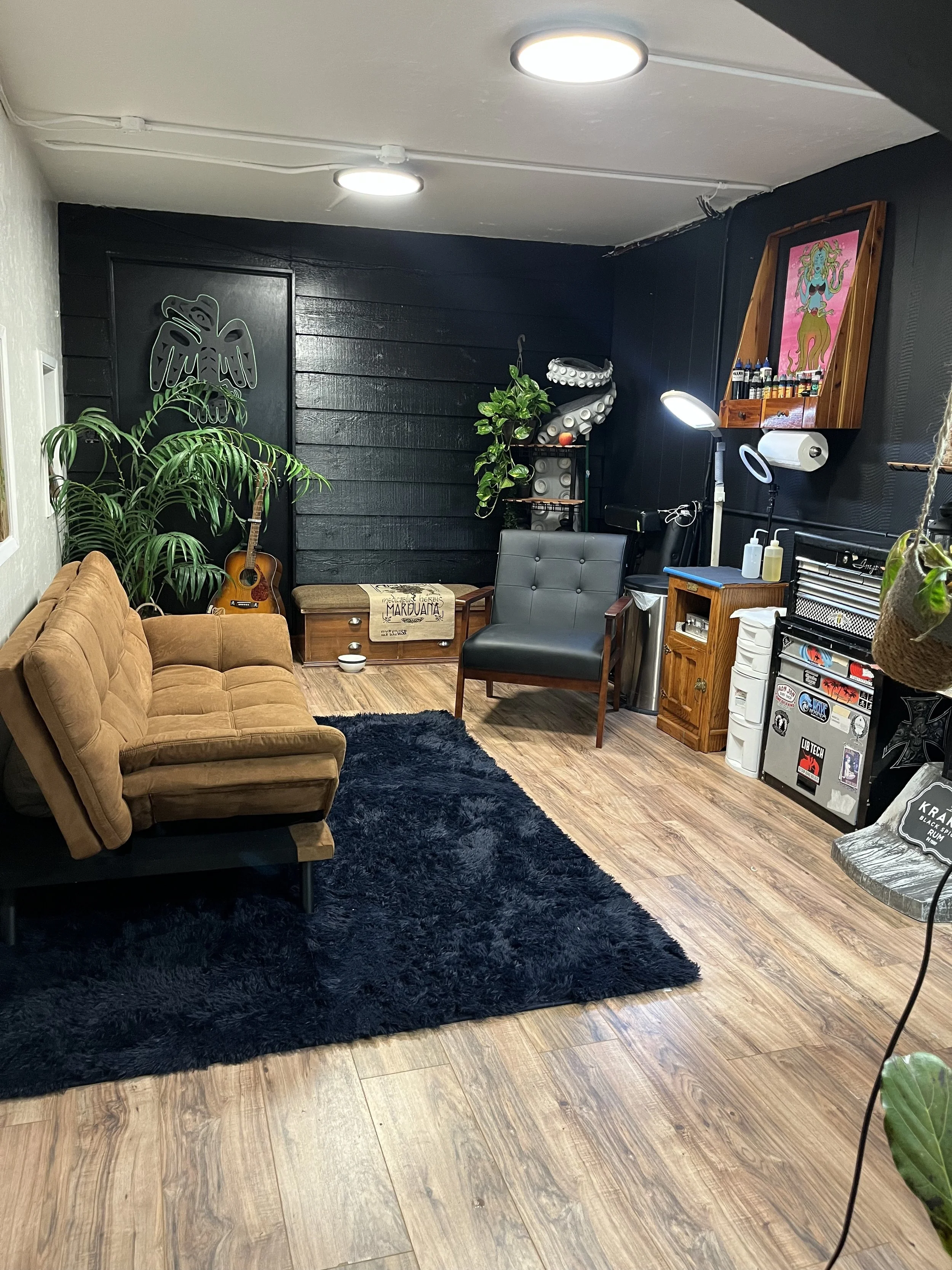 A cozy room with a tan sofa, a black shaggy rug, a green potted plant, a guitar, and eclectic artwork on the walls. There is a black wooden wall behind the guitar and some small furniture, including a wooden cabinet with a Marduata sign and a tattoo machine on a stand. The ceiling has two round lights and various plants and decorative items, creating a relaxed, artistic atmosphere.