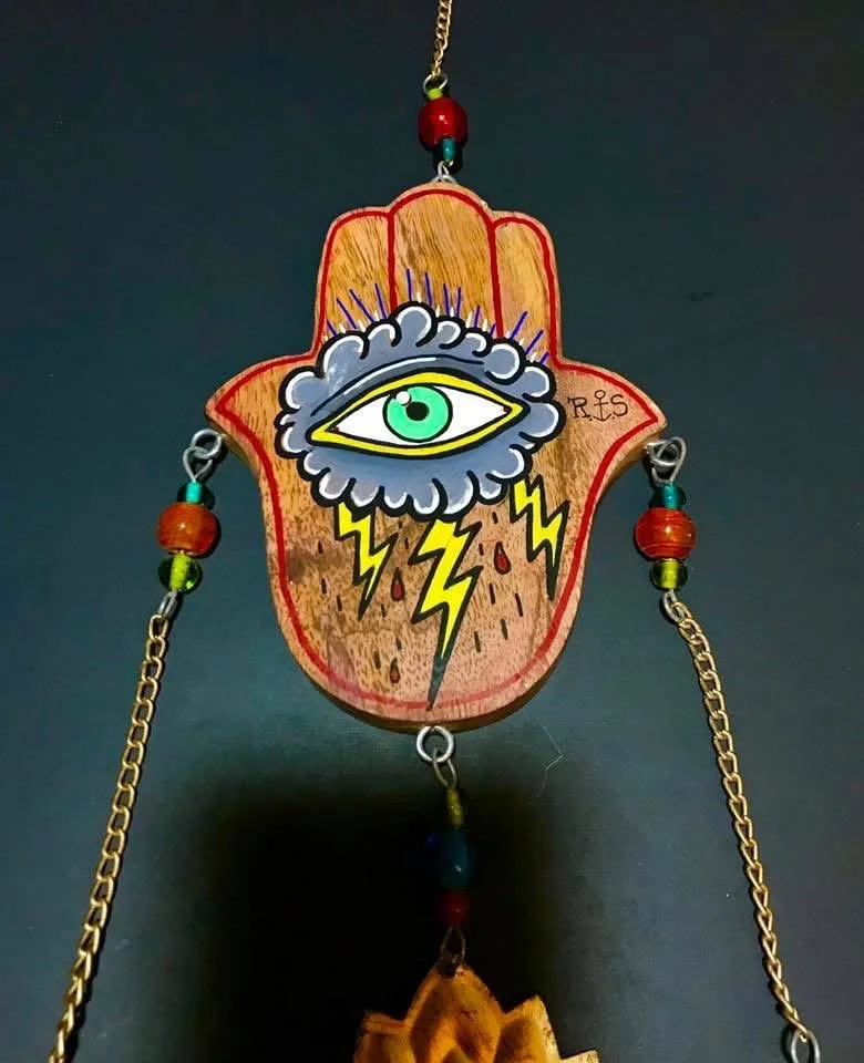 Decorative wall hanging featuring an illustrated eye with purple clouds, yellow lightning bolts, and Raindrop symbols, attached to a hand-shaped wooden background with beads and chains.