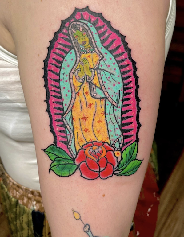 Colorful tattoo of Our Lady of Guadalupe with a red rose at the base and green leaves, within a pink and black border.