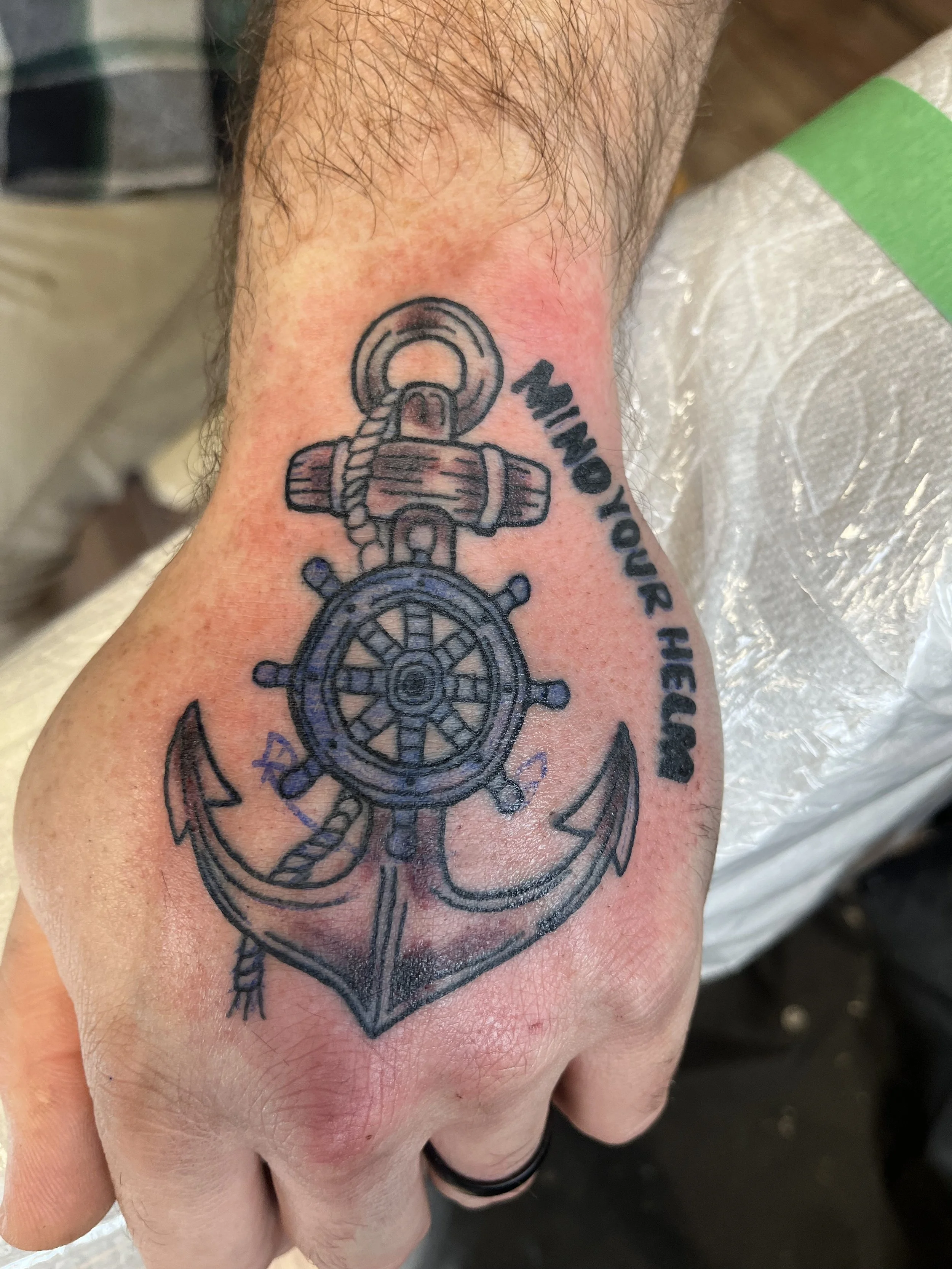 Tattoo of an anchor with a ship wheel and rope on the hand, with the phrase 'Who you hear him' written beside it.