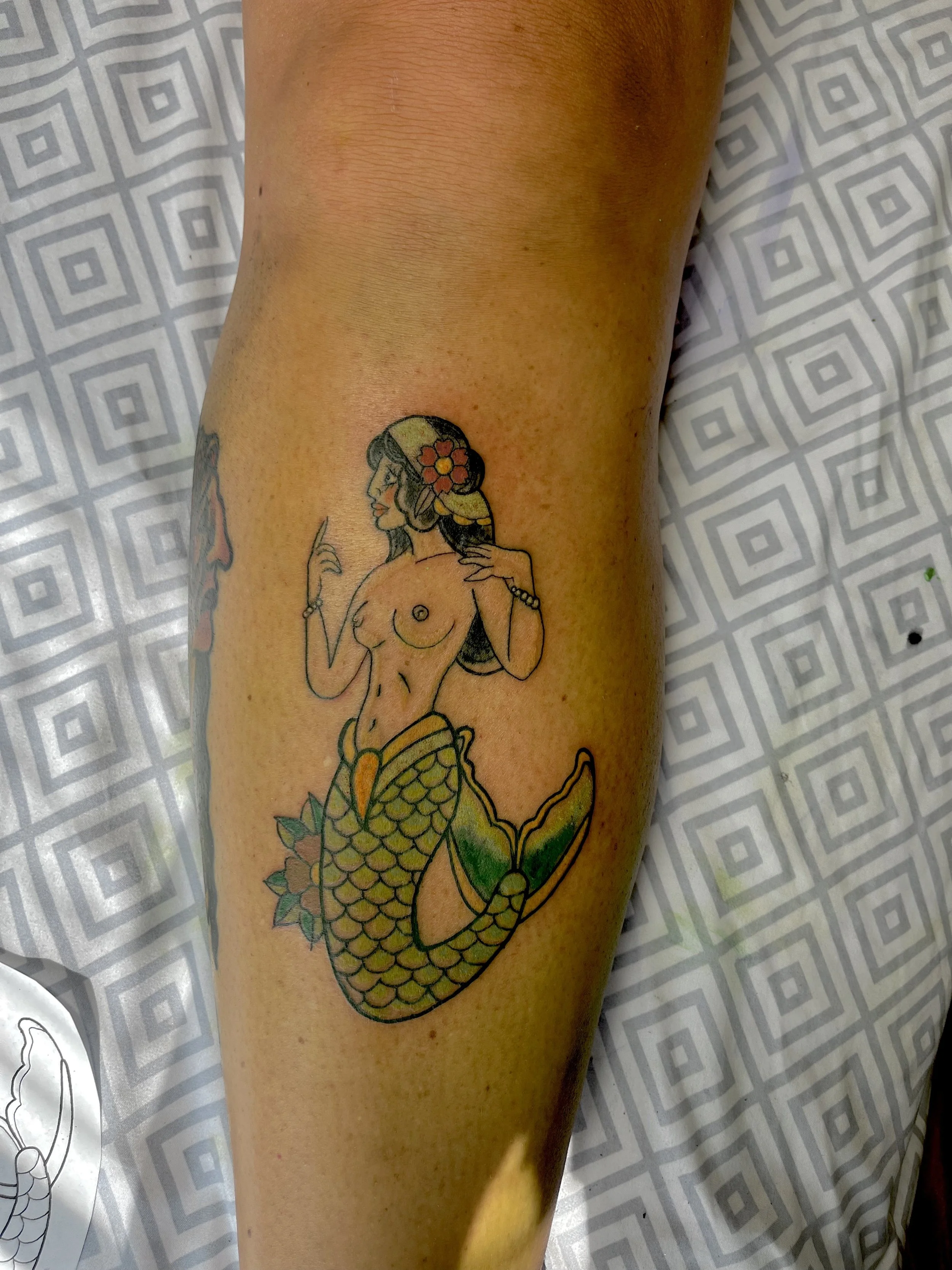 Tattoo of a mermaid with a flower in her hair on a person's forearm.