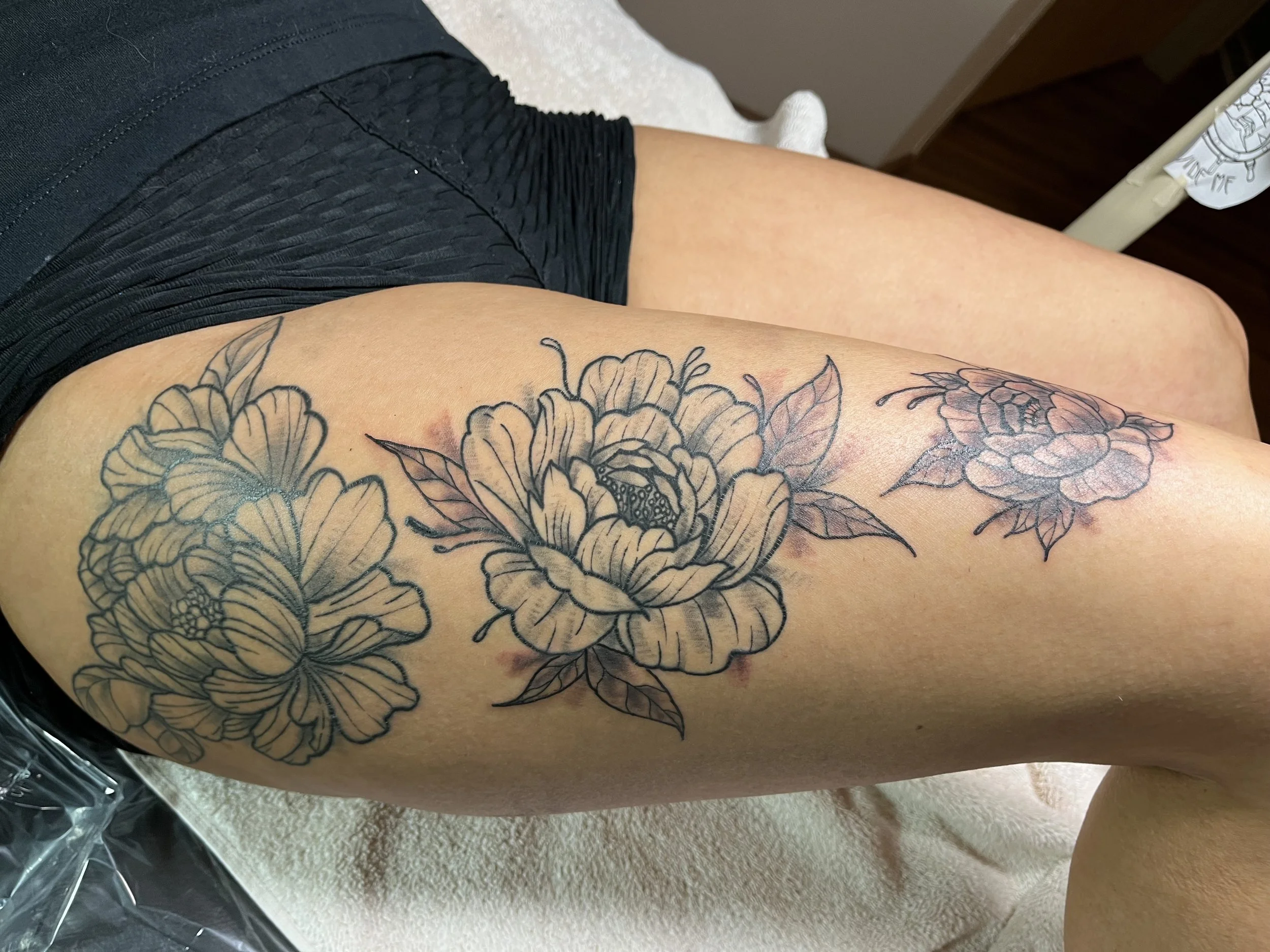 A person's thigh with a floral tattoo consisting of three large flowers with detailed petals and leaves.