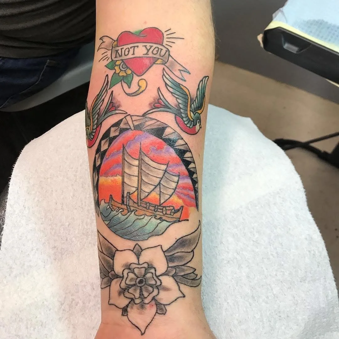 Tattoo on forearm featuring a flying swallow, a heart with a banner that says 'NOT YOU', a sailing ship against a sunset sky, and a flower at the wrist.