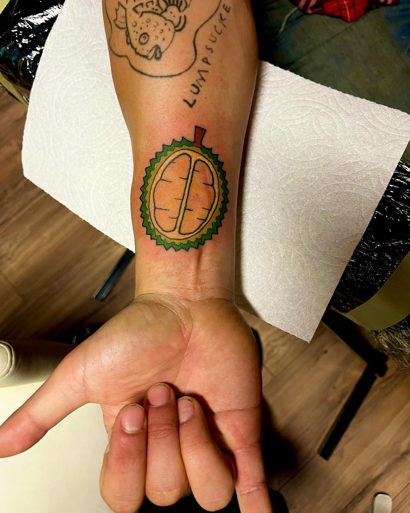 Colorful tattoo of a durian fruit on person's forearm, with a cartoonish style and 'LUMP SUCK' written above.