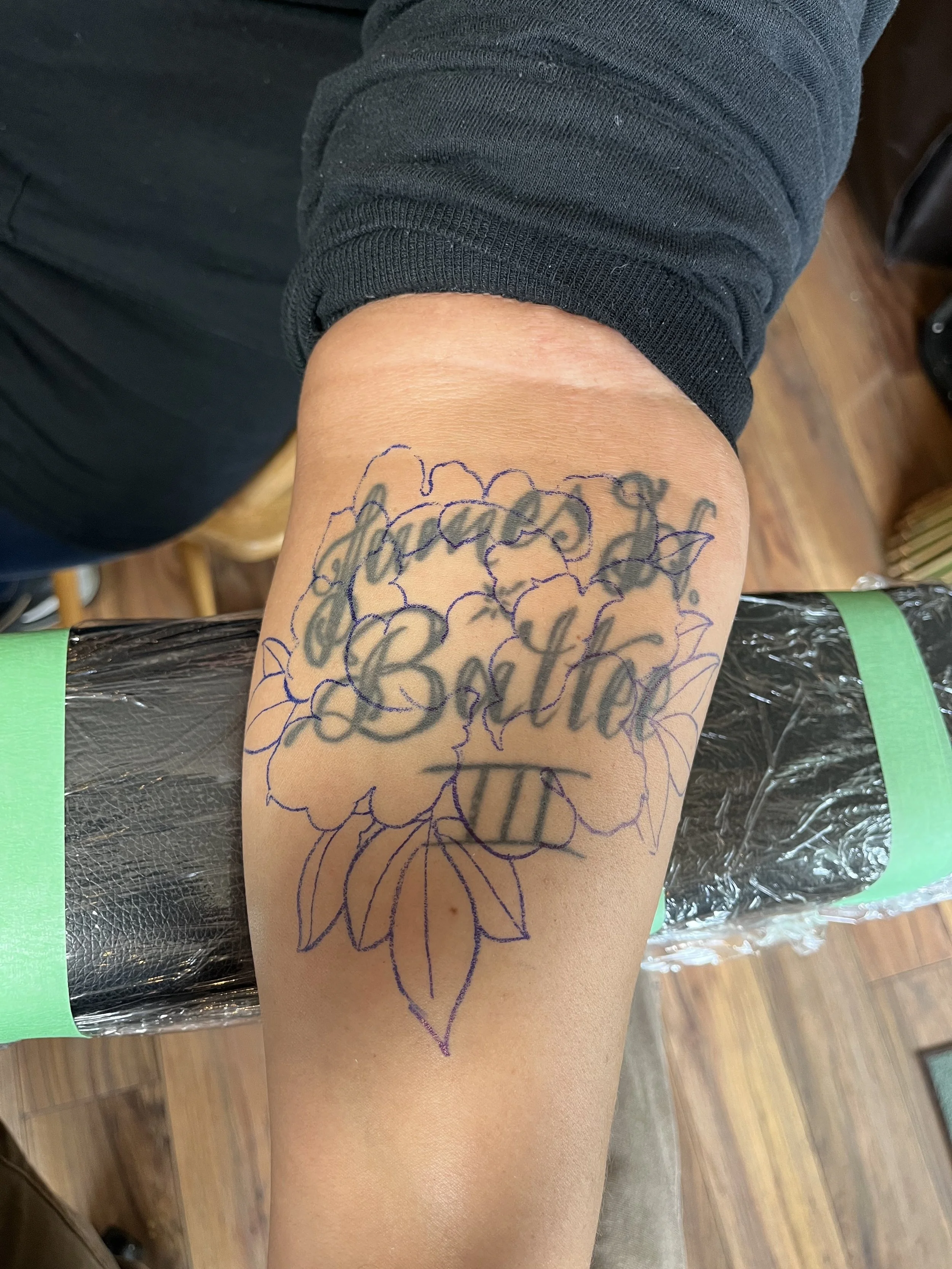 A tattoo stencil of large flowers with the words 'Grandma' and 'Bata' overlaid in script, on a person's upper arm.