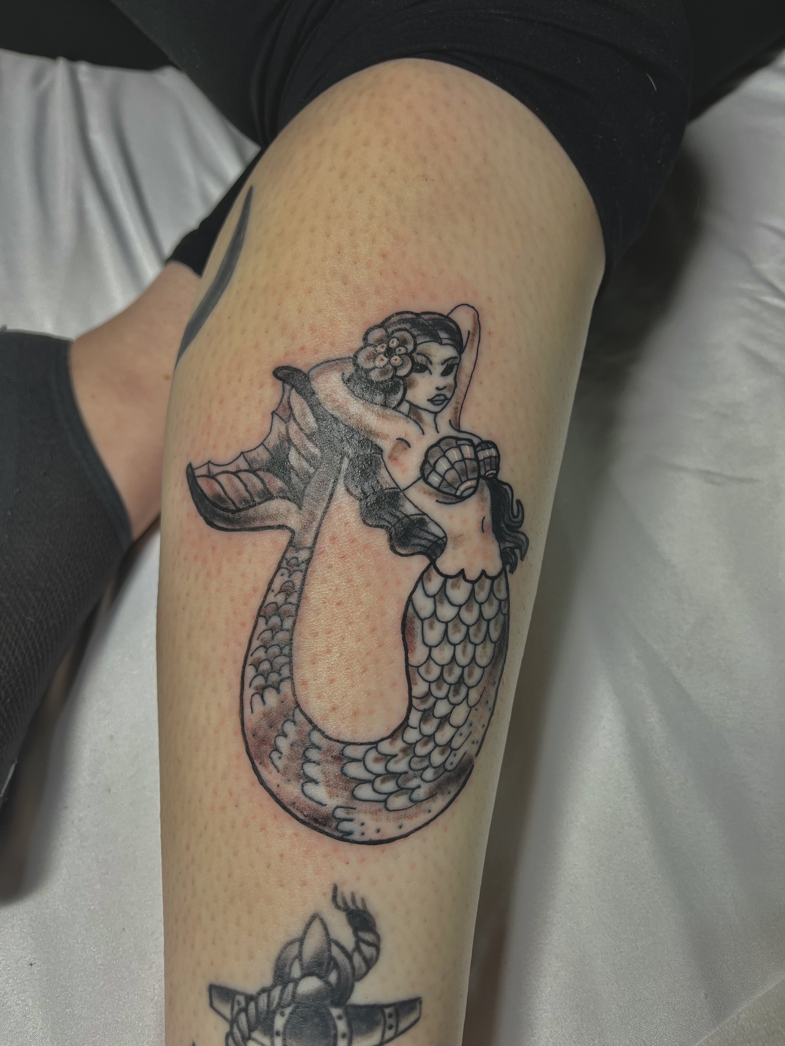 Tattoo of a mermaid with long hair and flower accessories, holding a sword, with a fish tail, on a person's thigh.
