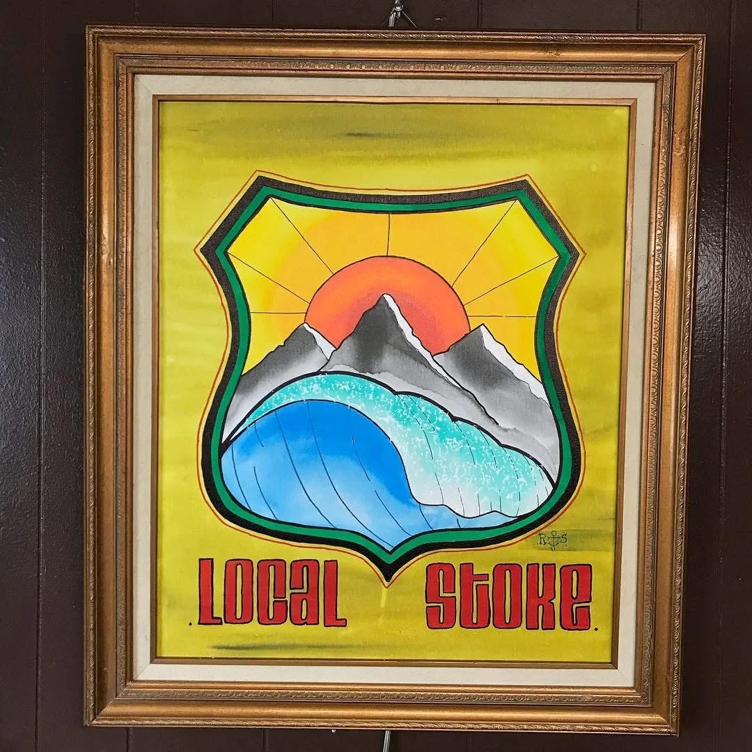 A colorful framed painting with a yellow background, depicting mountains, a waterfall, and the sun, with the words "Local Stoke" written in red at the bottom.