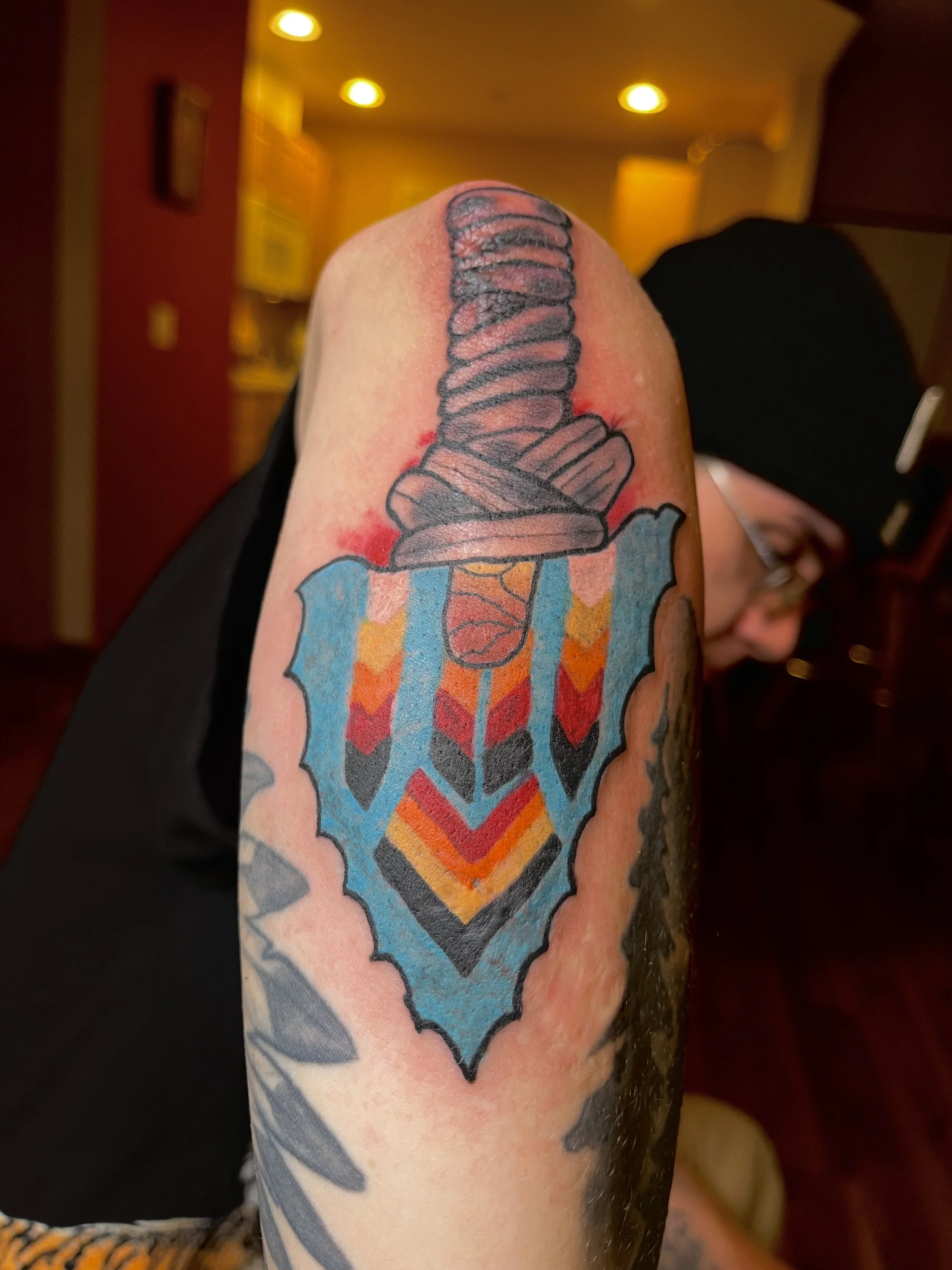 Tattoo of a colorful arrow pointing downward with a stylized arrowhead, on a person's arm.
