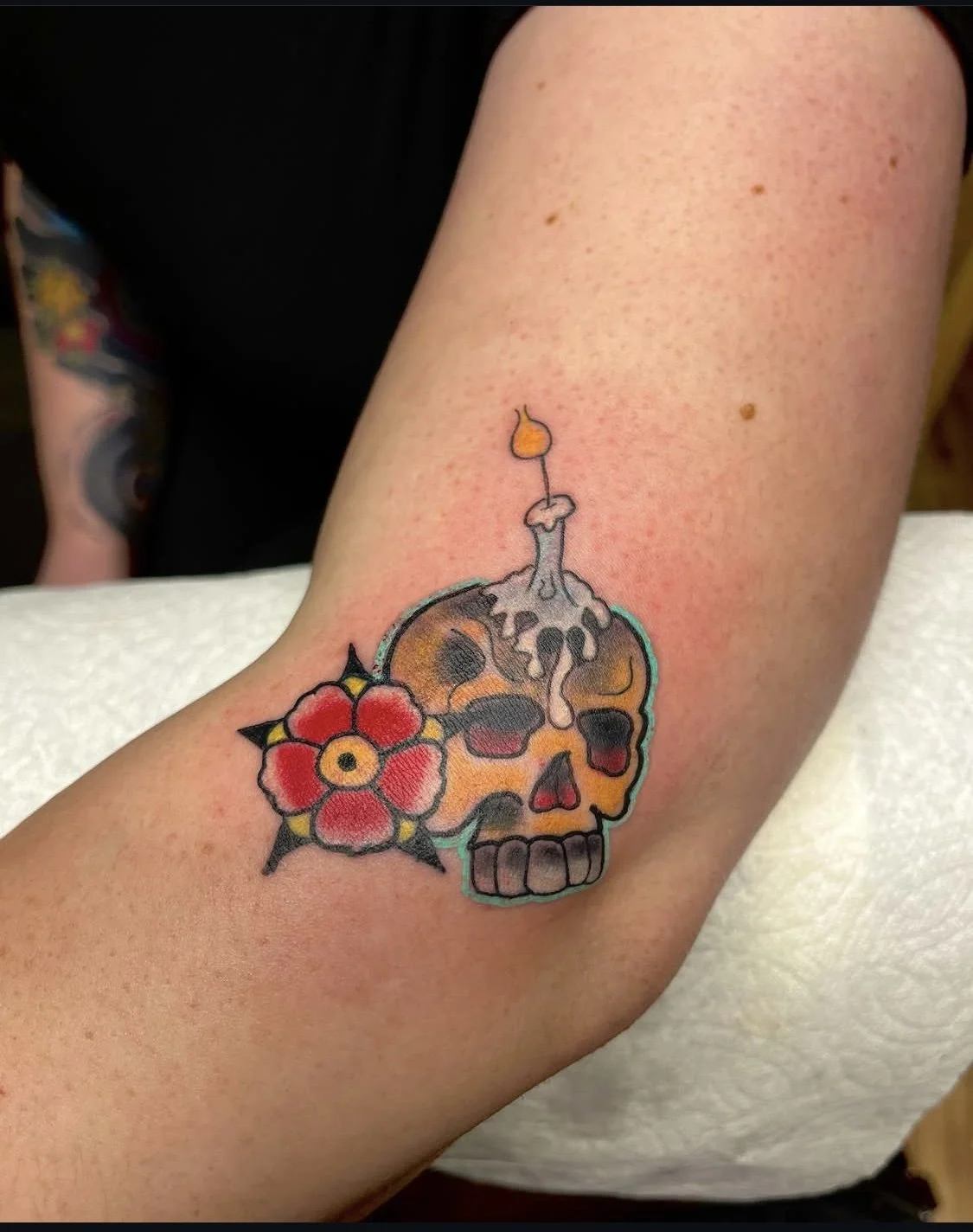Tattoo of a skull with a candle on top, surrounded by a red flower with a yellow center and black outlines, on a person's forearm.