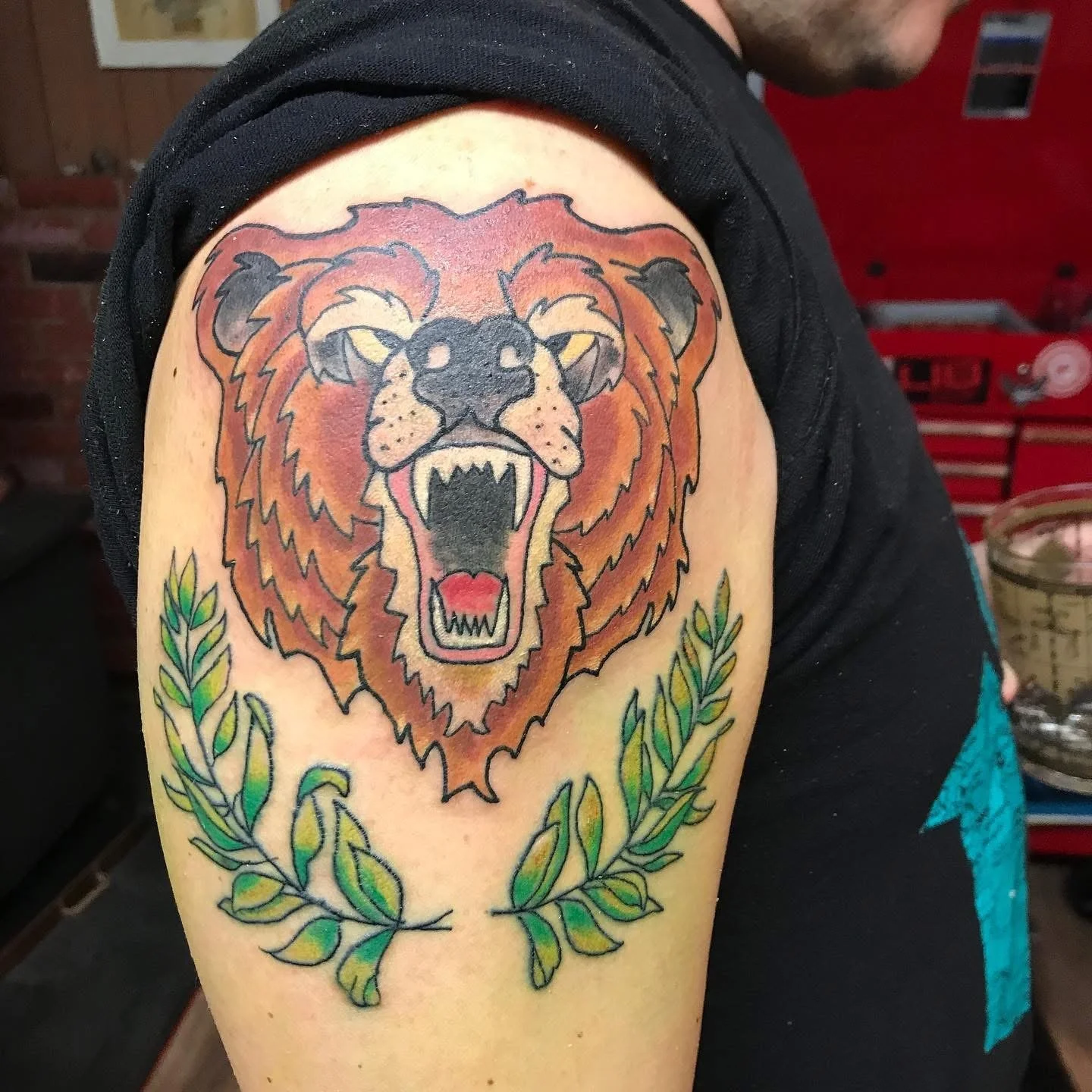 Tattoo of a roaring lion's head with a black nose, sharp teeth, and a red tongue, surrounded by green laurel branches on a person's upper arm.
