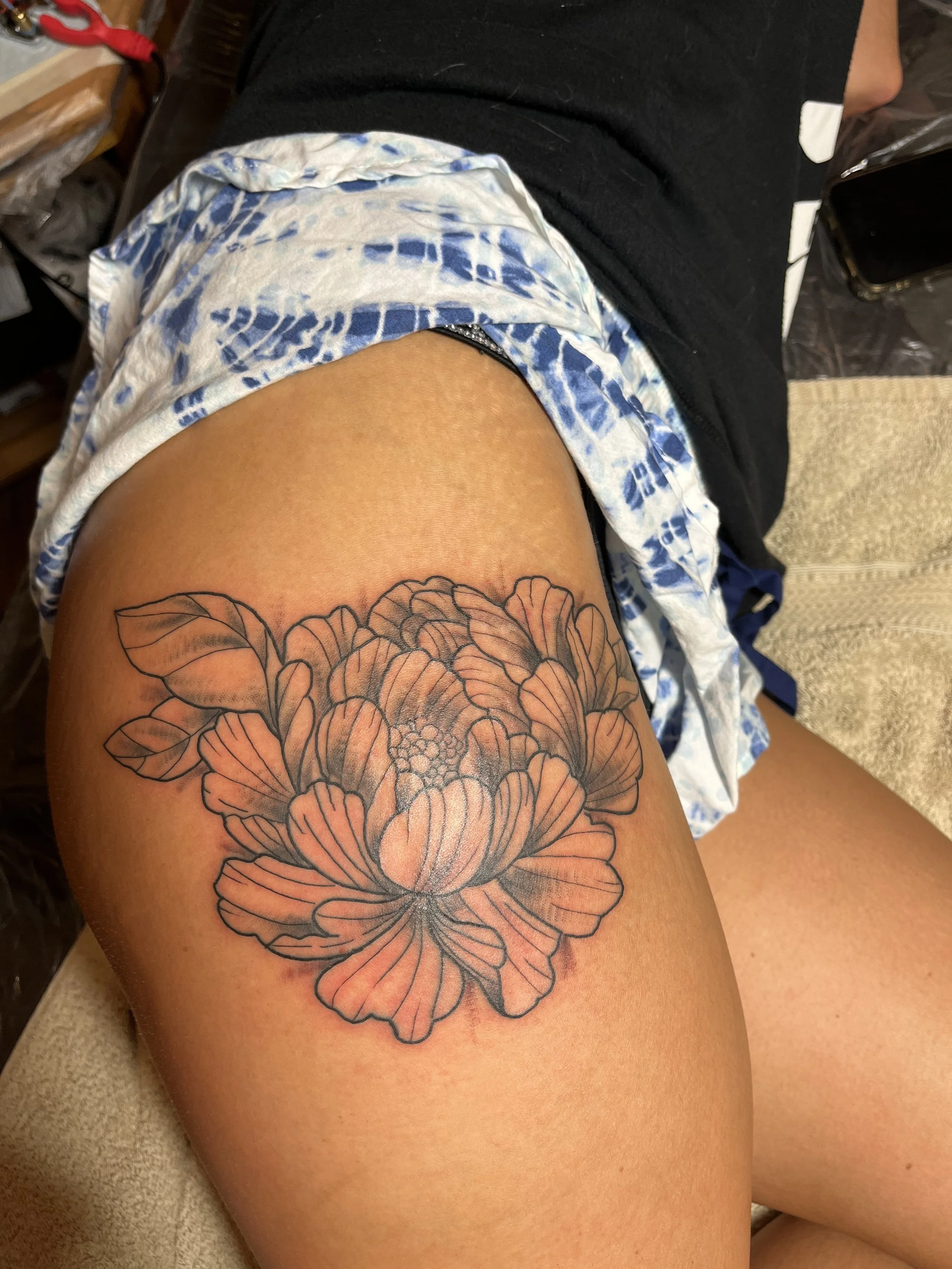 A person's thigh with a large black outline floral tattoo of a peony. The tattoo features shading in the petals, and the person is wearing a blue and white tie-dye shirt with black shorts.
