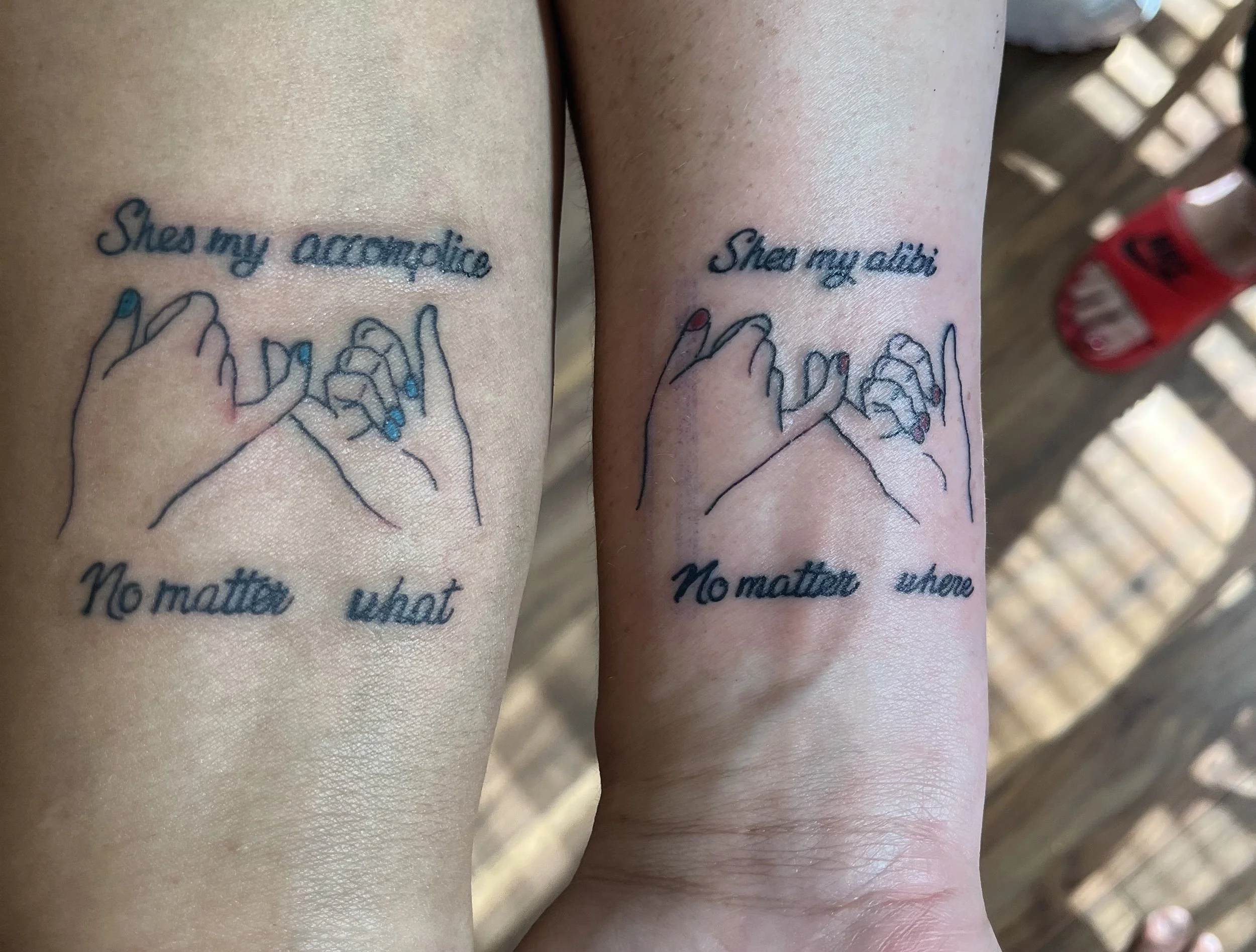 Two wrists with matching tattoos of hands holding each other, with the text "She's my accomplice" above and "No matter where" below.