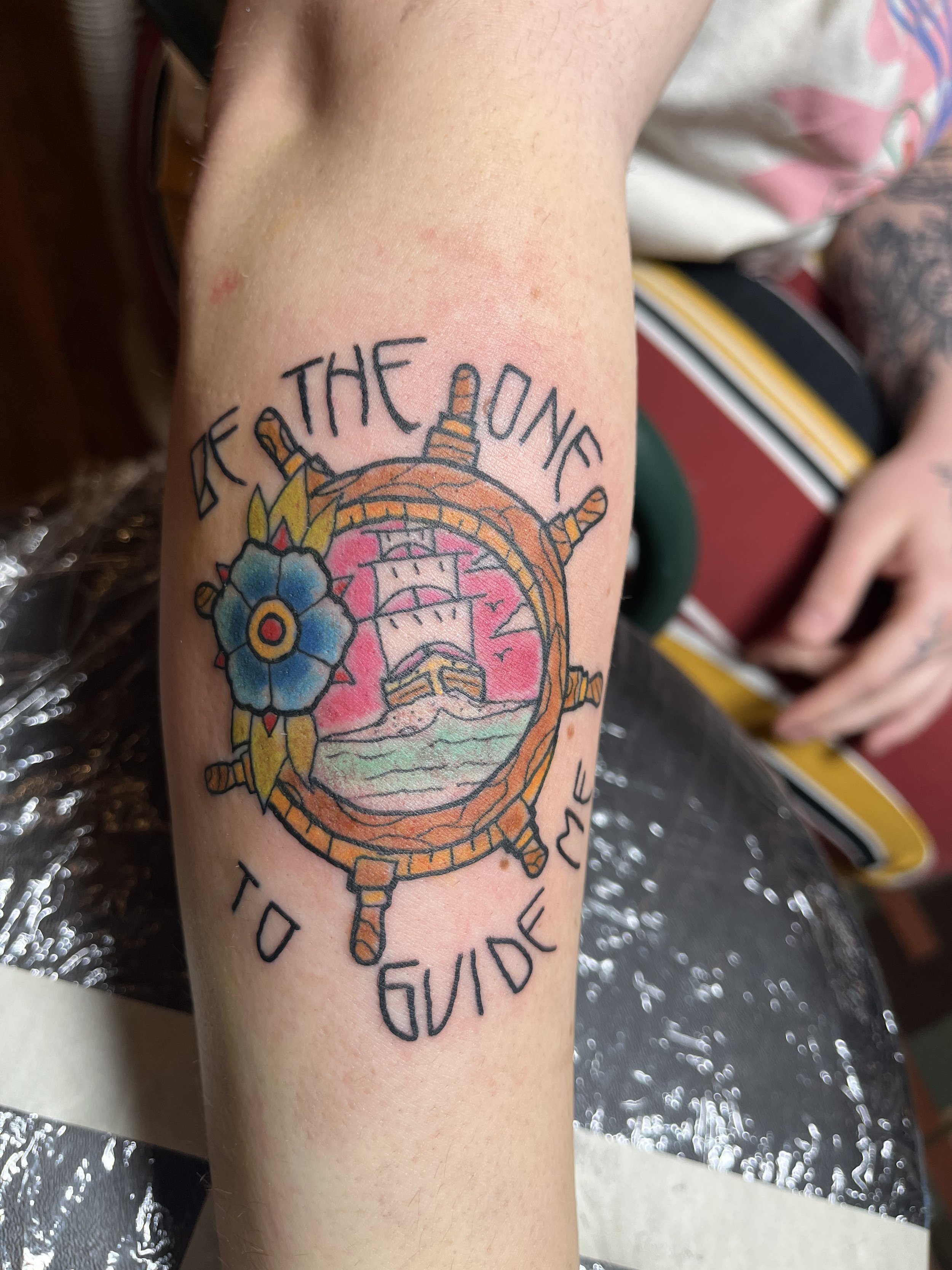 Tattoo of a ship inside a ship's wheel with a pink sunset, a blue flower, and the words 'Set the course to guide me'.
