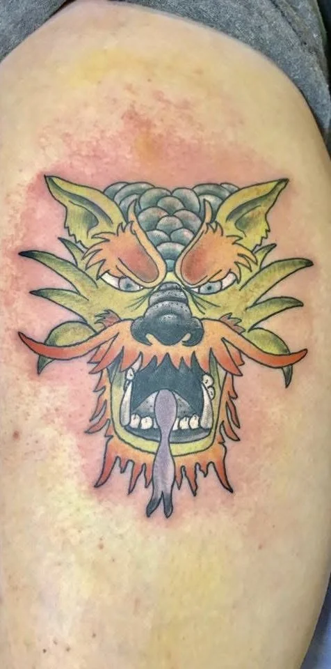 Colorful tattoo of a fierce dragon's head with sharp eyes, open mouth showing teeth, and a long tongue, on a person's skin.