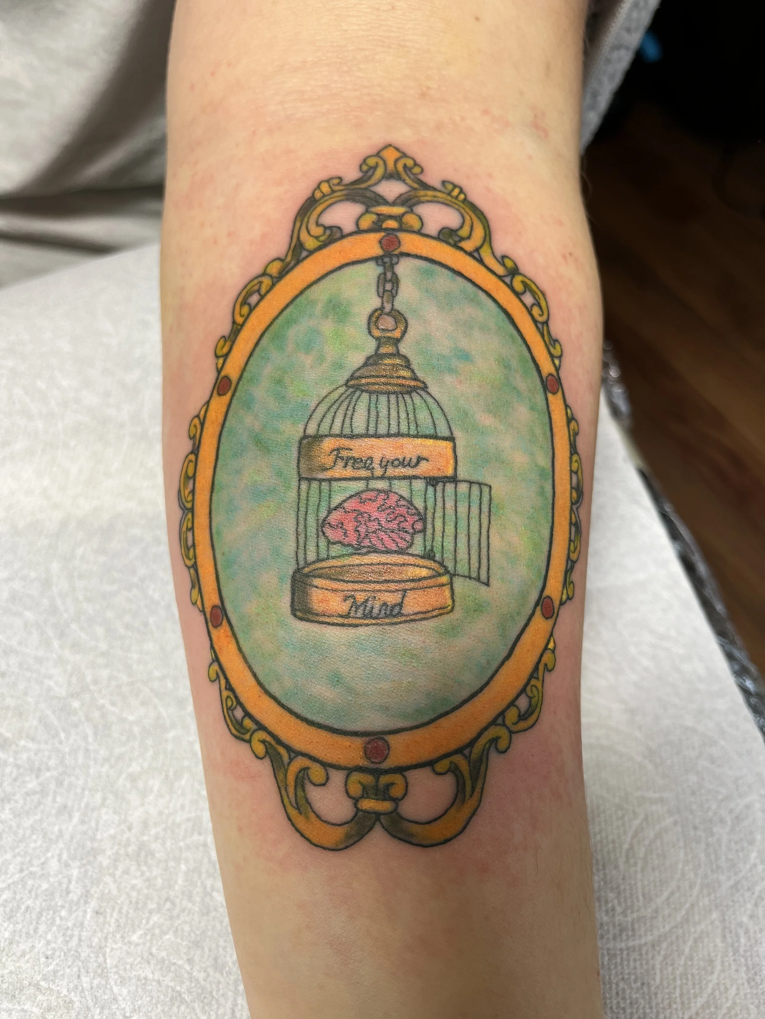 Tattoo of a birdcage with a brain inside, framed by an ornate oval border.