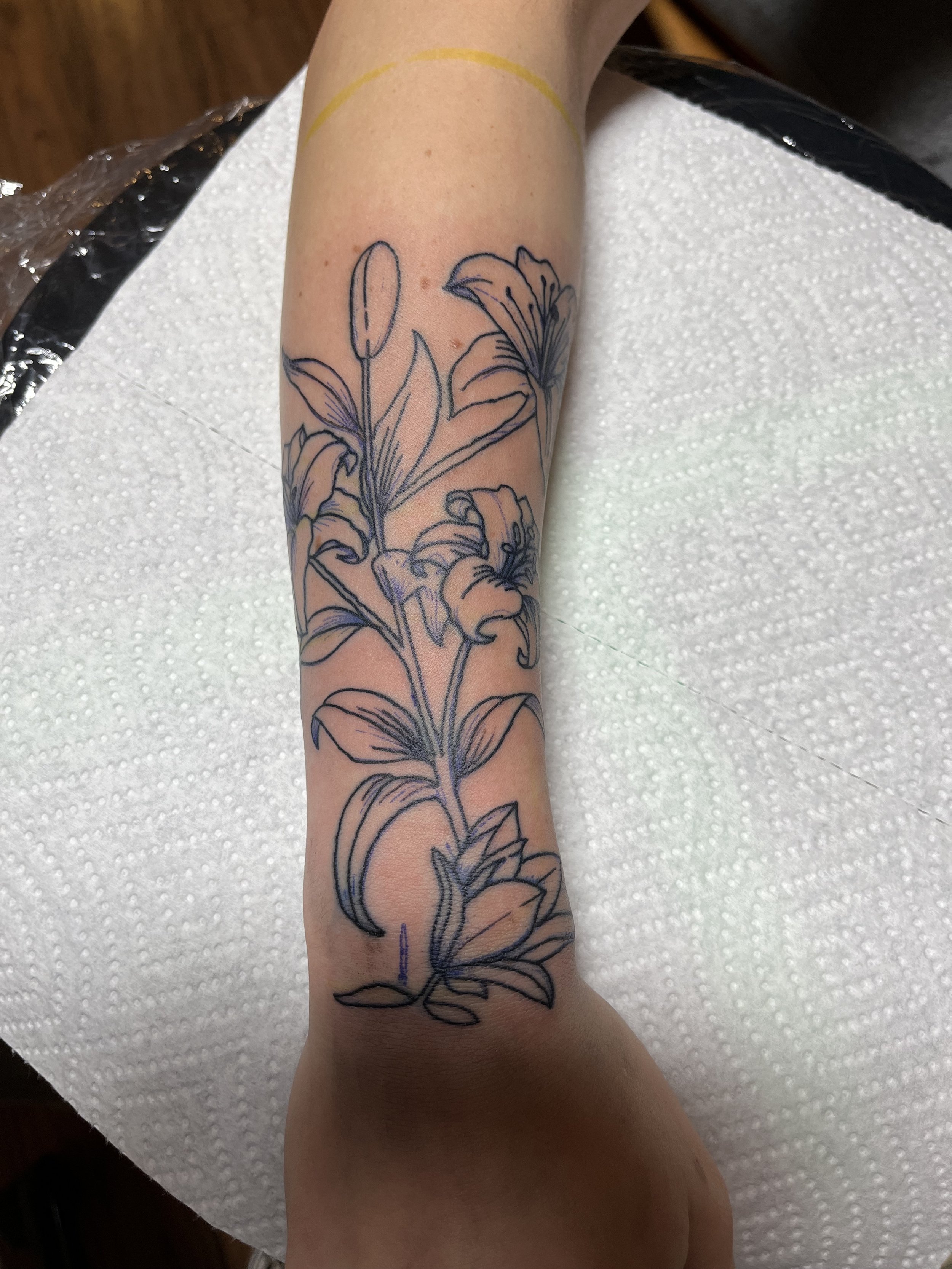 Black outline tattoo of a floral design on a person's forearm with lilies and leaves.
