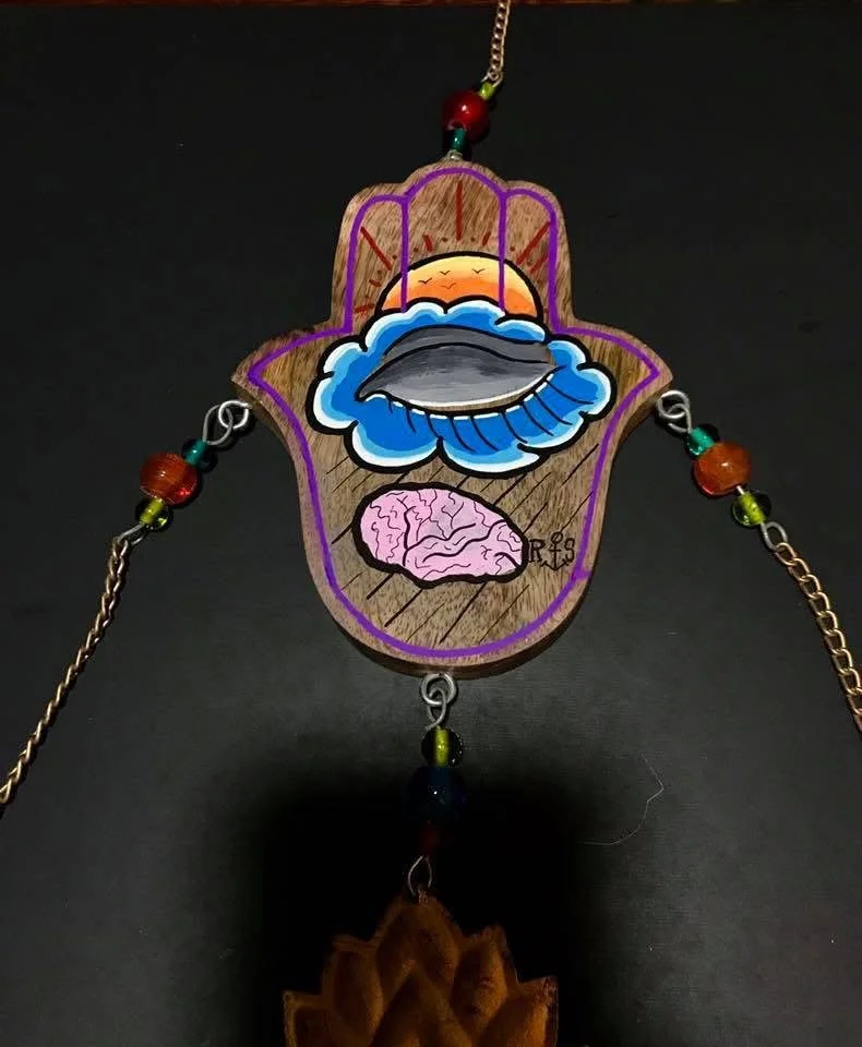 An artistic necklace featuring a wooden pendant with a painted scene of a sunset, clouds, and a brain, decorated with colorful beads and chains.