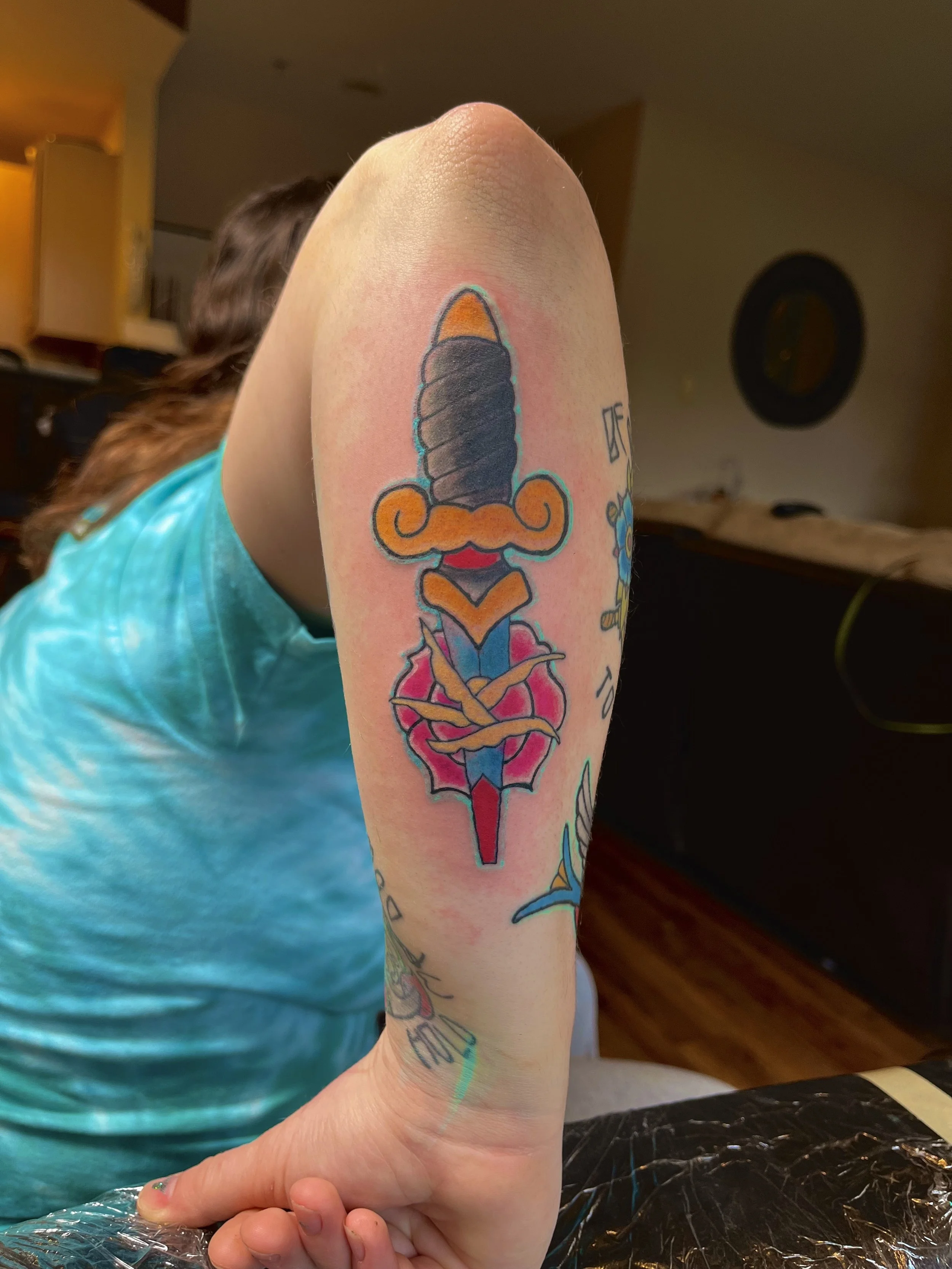 Tattoo of a colorful dagger with a black blade, orange curly flourish, pink and blue leaves, and a red tip, on a person's upper arm.