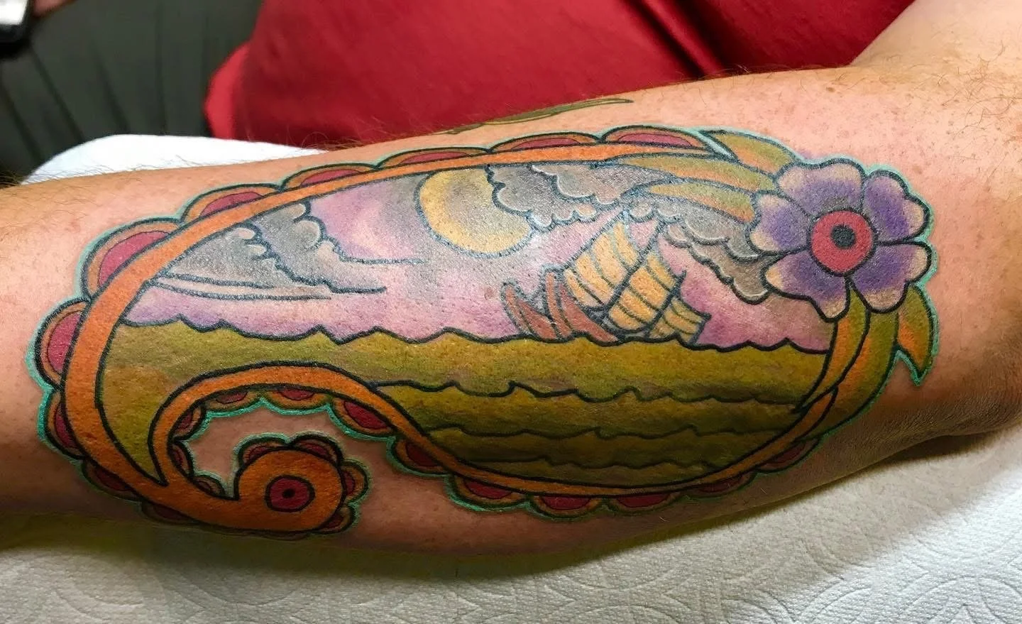 Colorful tattoo of a seashell with a ship, clouds, moon, and flowers on a person's arm.
