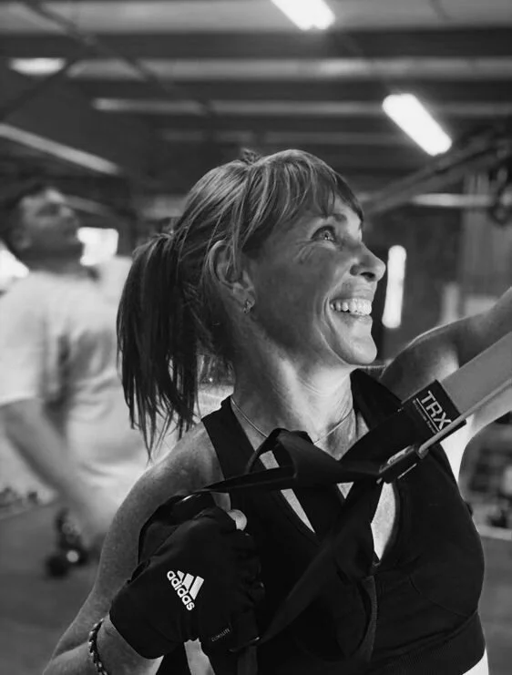 Close-up of a woman smiling after workout in gym, wearing gloves and sports attire, with a man blurred in the background.