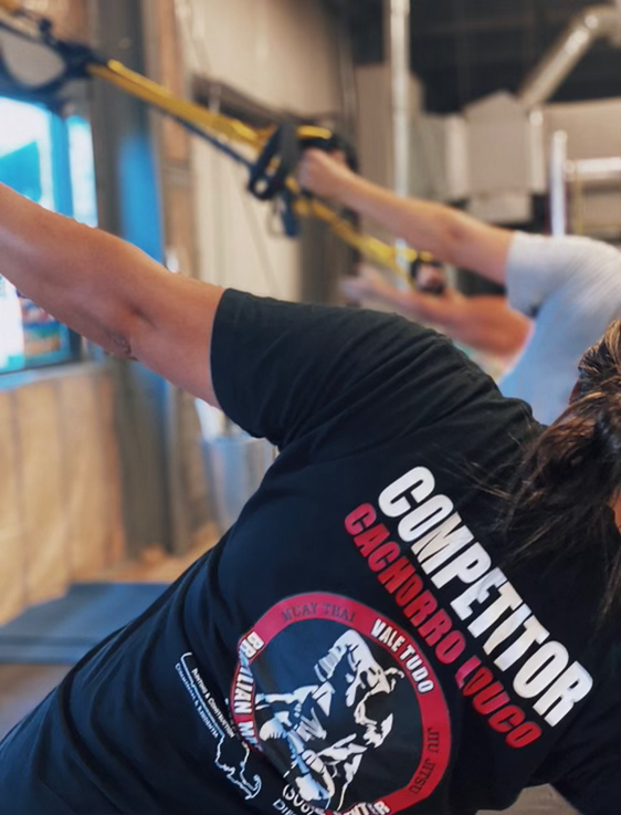 A woman performing a resistance training exercise with a yellow strap in a gym. The person is wearing a black T-shirt with text and graphics related to combat sports.