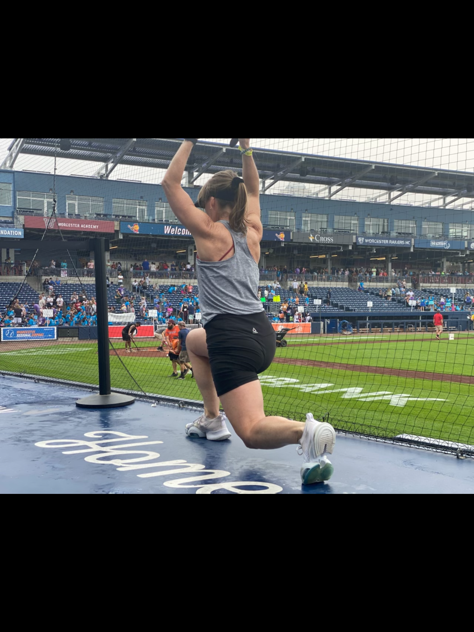 Female athlete leading stretching activities on the sidelines of a Polar Park, wearing a gray tank top and black shorts, with a baseball stadium and spectators in the background.