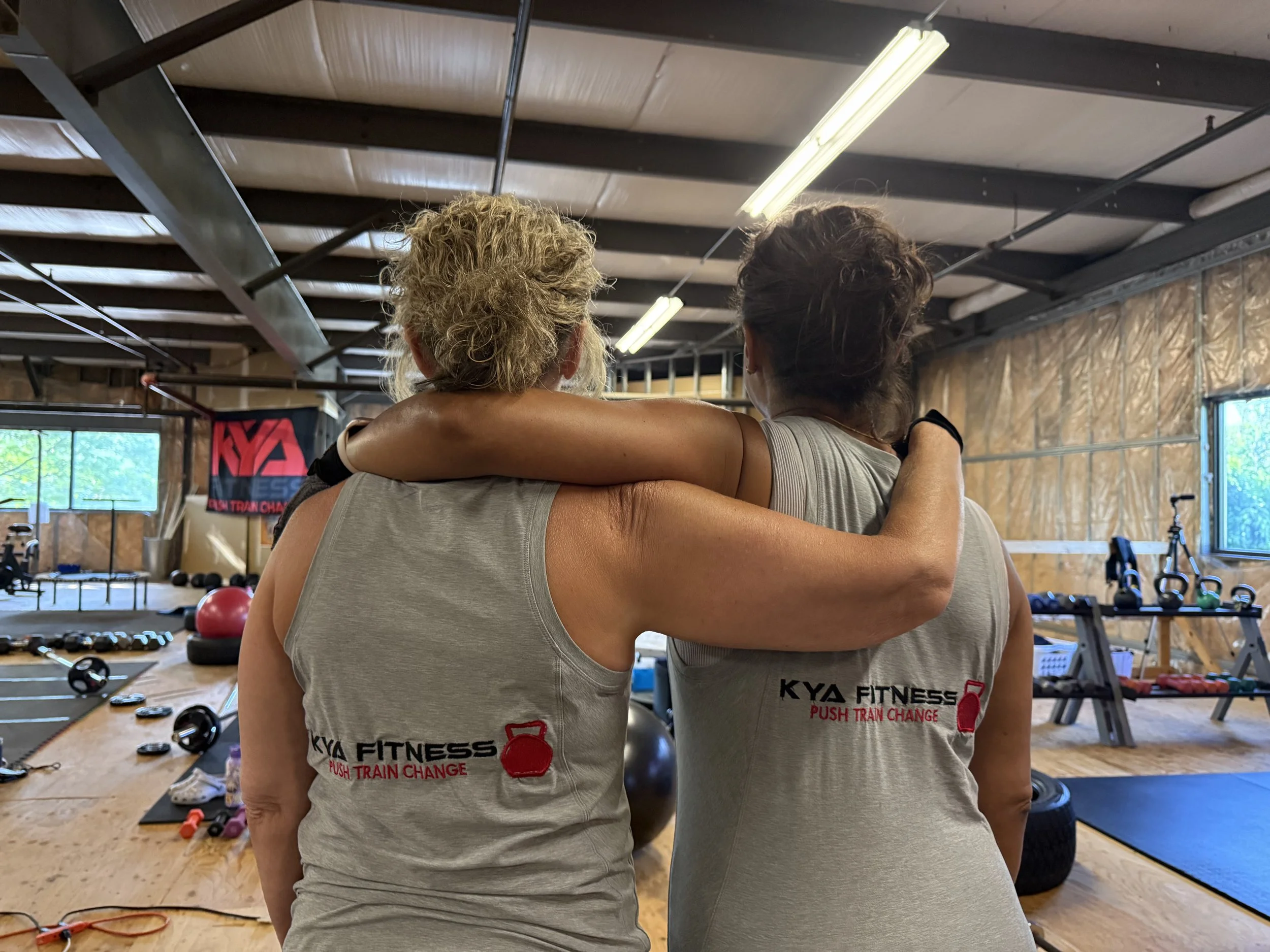 Two women in gray workout tank tops with 'KYA FITNESS' and 'PUSH TRAIN CHANGE' logos embrace each other in a gym, seen from behind, with gym equipment and a Bayern Munich flag in the background.
