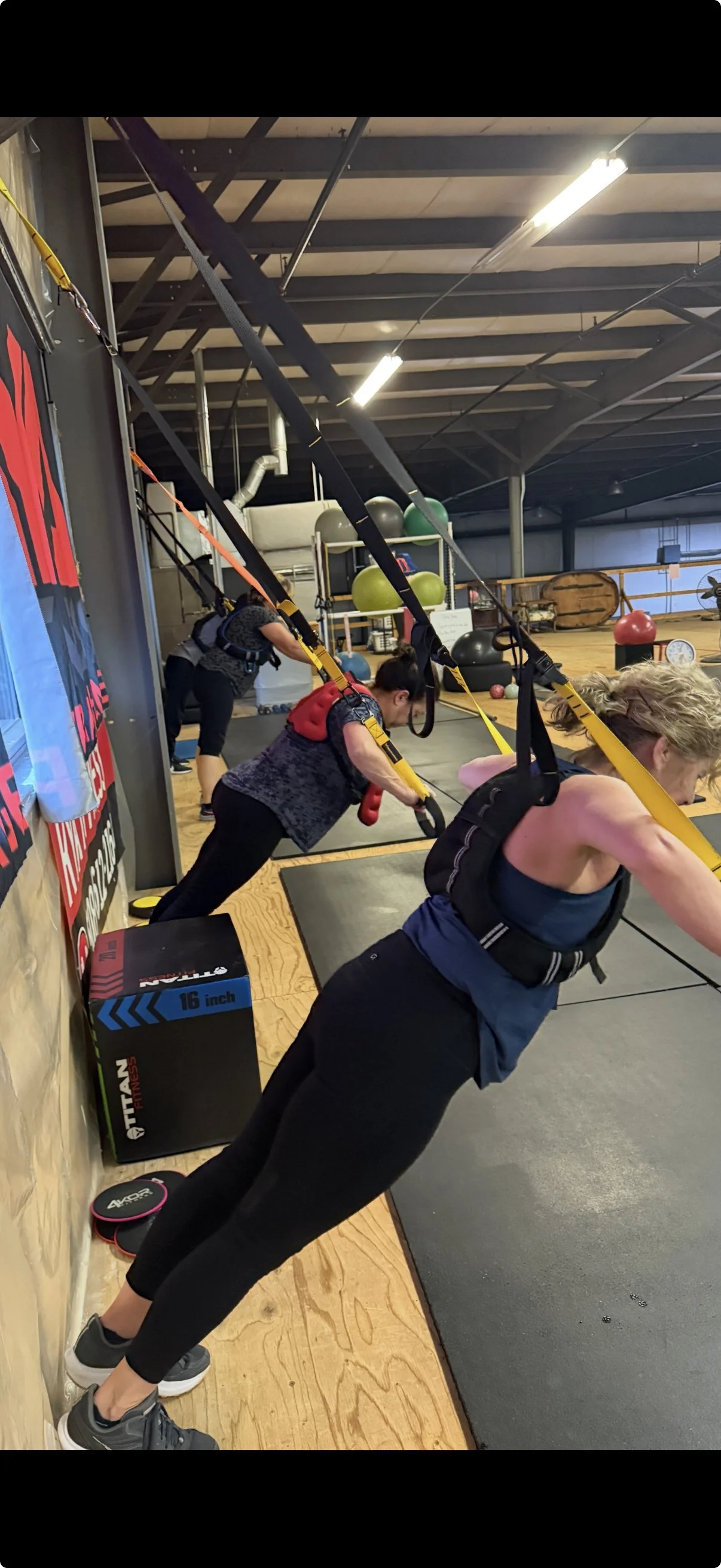 Women participating in TRX suspension training class leaning forward while holding TRX handles with their feet on the ground in a gym.