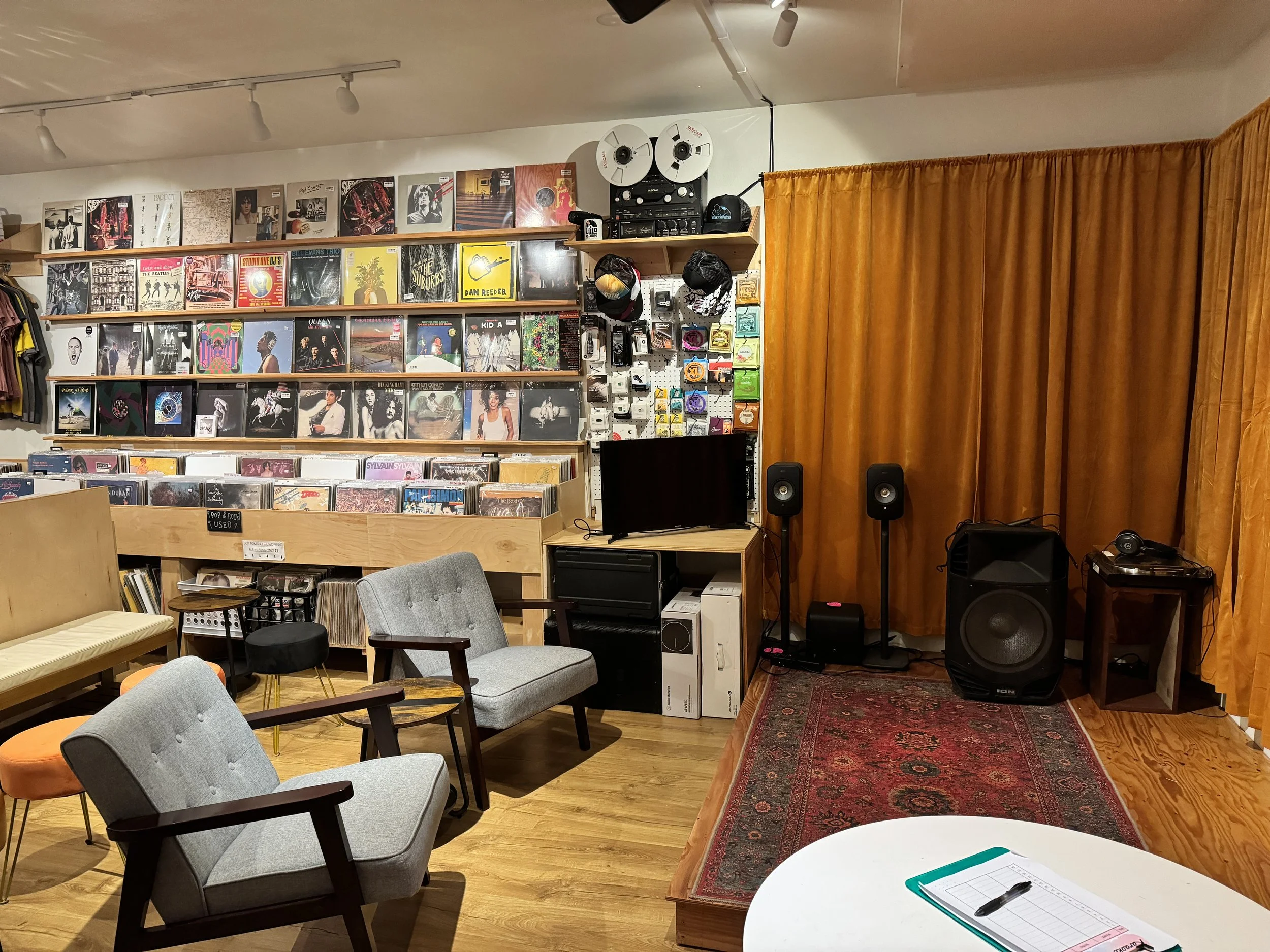 Interior of a cozy music store or cafe with vinyl records on shelves, orange curtains, wooden flooring, cushioned chairs, and a small stage area with audio equipment and speakers.