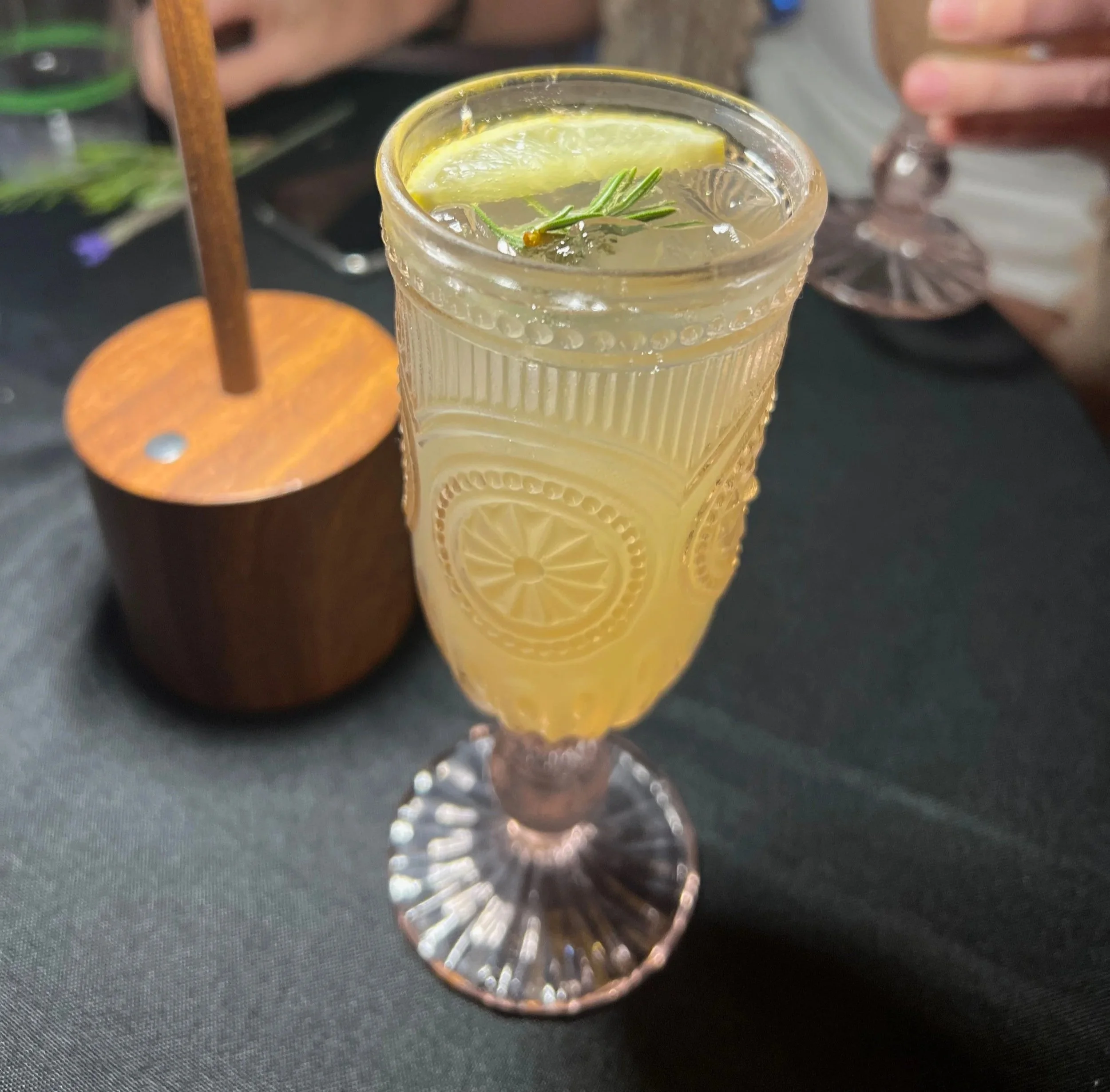A tall, decorative glass of lemonade garnished with a lemon wedge and a sprig of rosemary, on a black table.