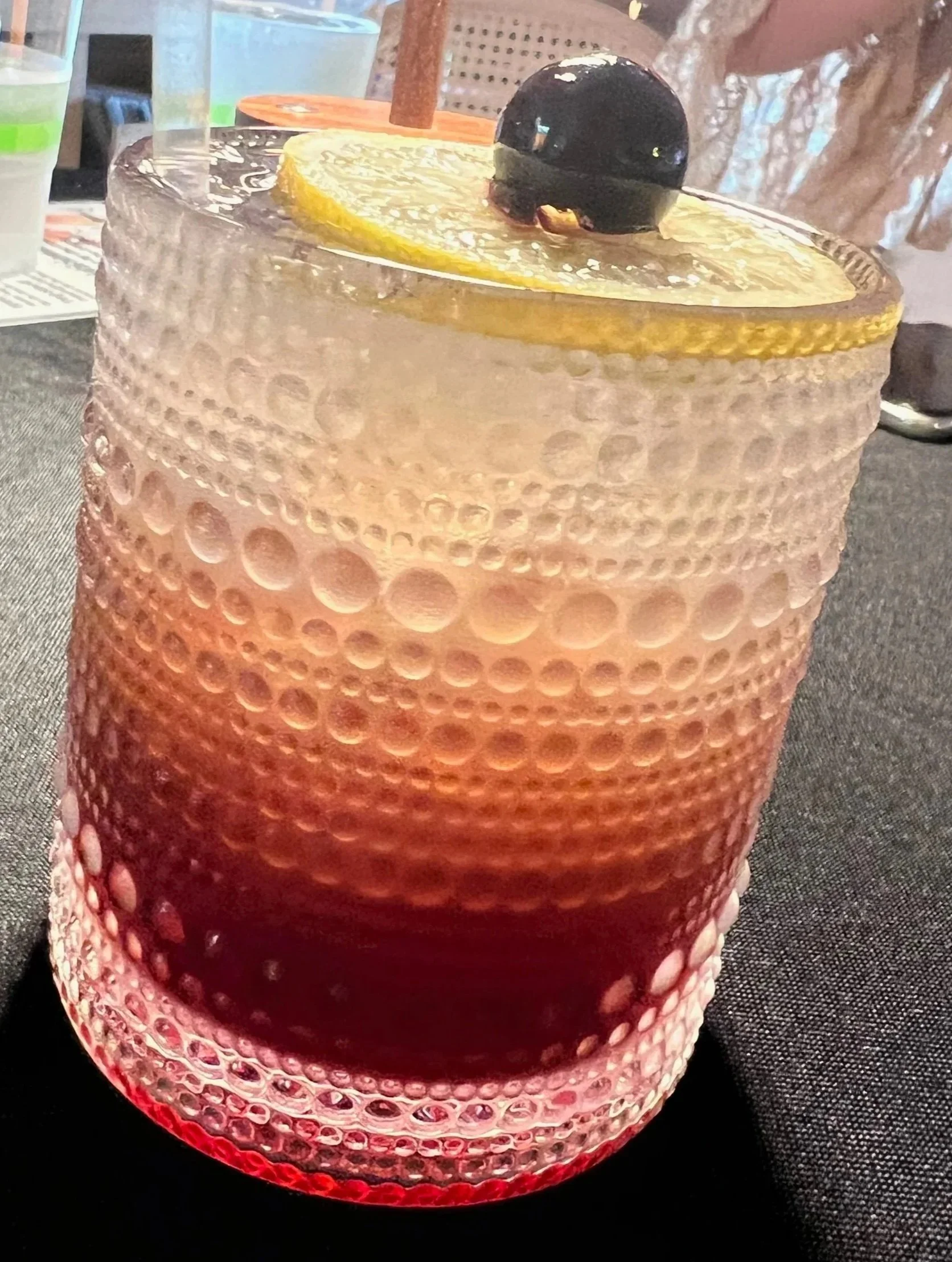 A textured glass containing a red-colored cocktail with a lemon slice and a cherry garnish.