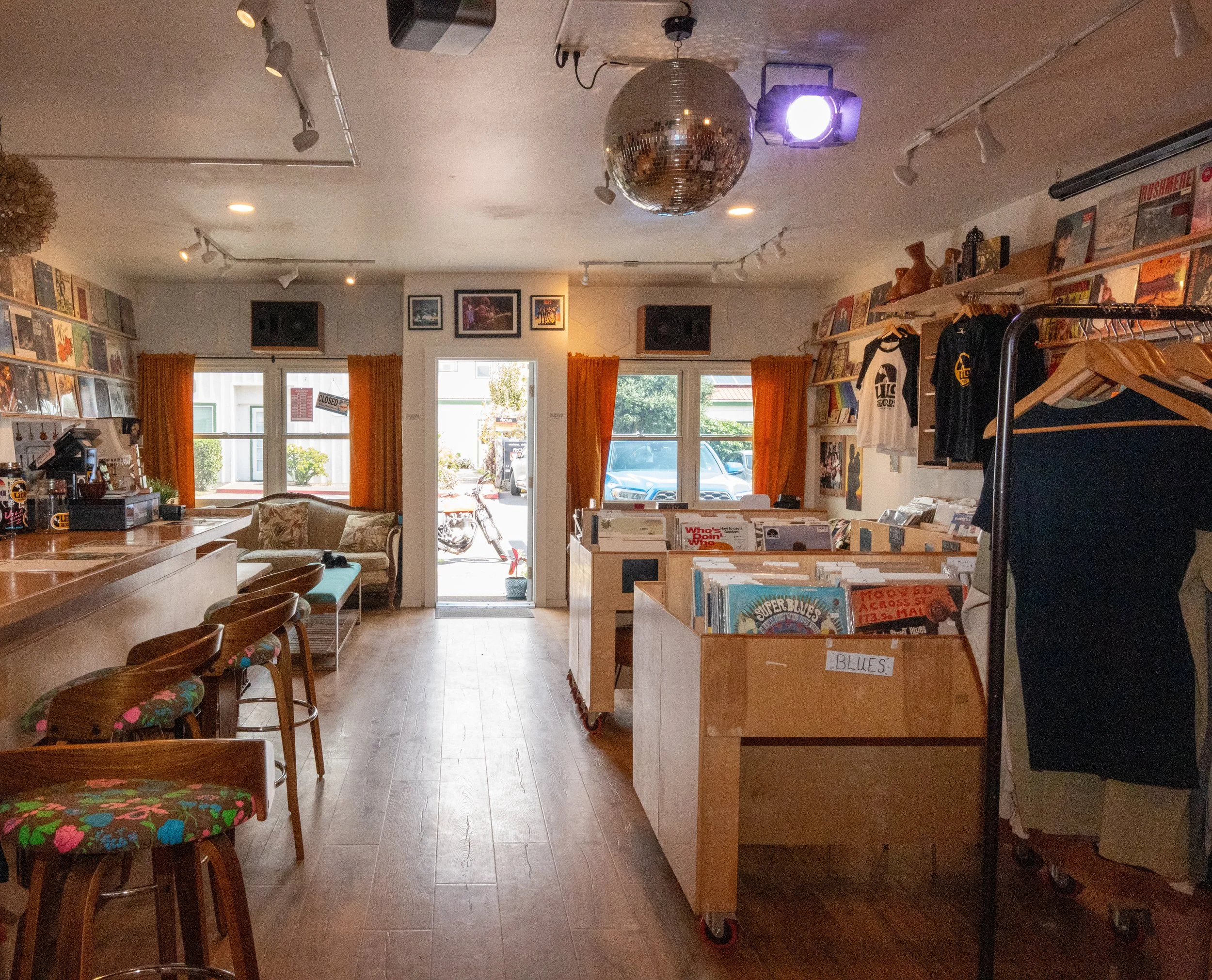 Inside a record store with wooden floors, orange curtains, vinyl records on shelves, a clothing rack, and a retro sofa near the door, illuminated by track lighting and a disco ball hanging from the ceiling.