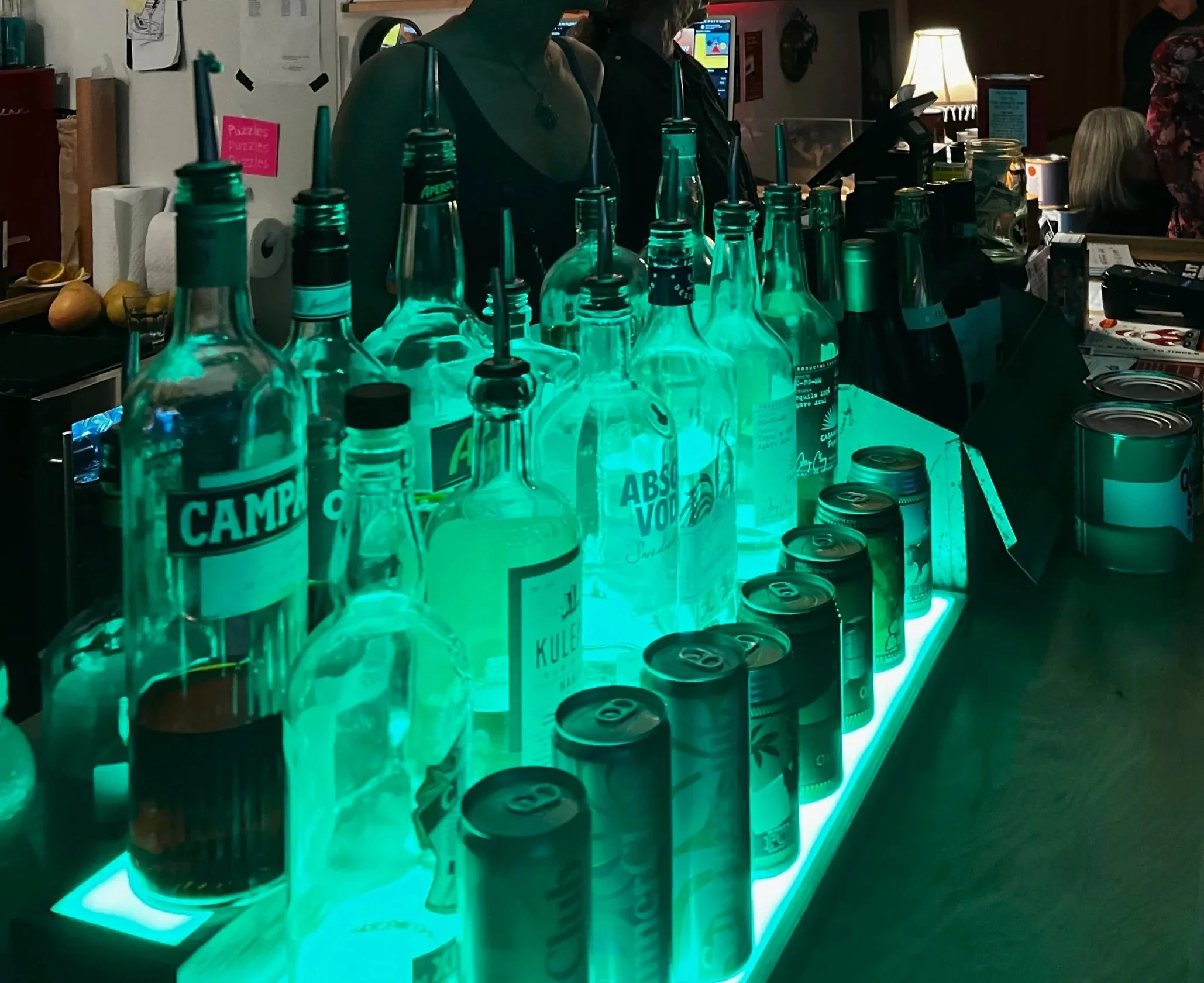 A bar with a glowing teal display of various alcohol bottles and cans, including brands like Campari and Absolut Vodka, with some people in the background.