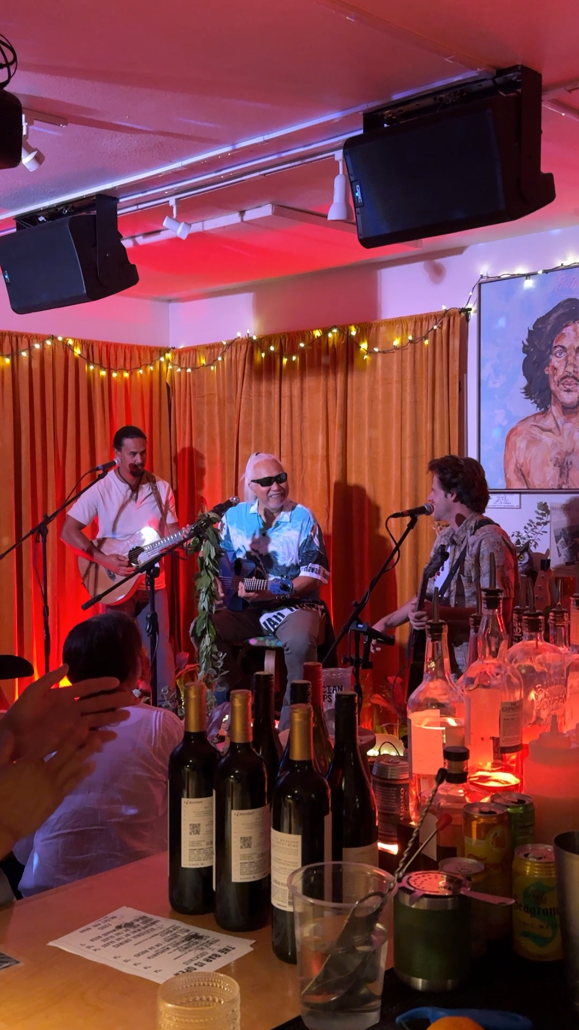 A live music performance at a cozy indoor venue with three male musicians playing guitars on stage; one with sunglasses, a large portrait painting of a man with curly hair on the wall, warm string lights, bottles and drinks on a table in the foreground.