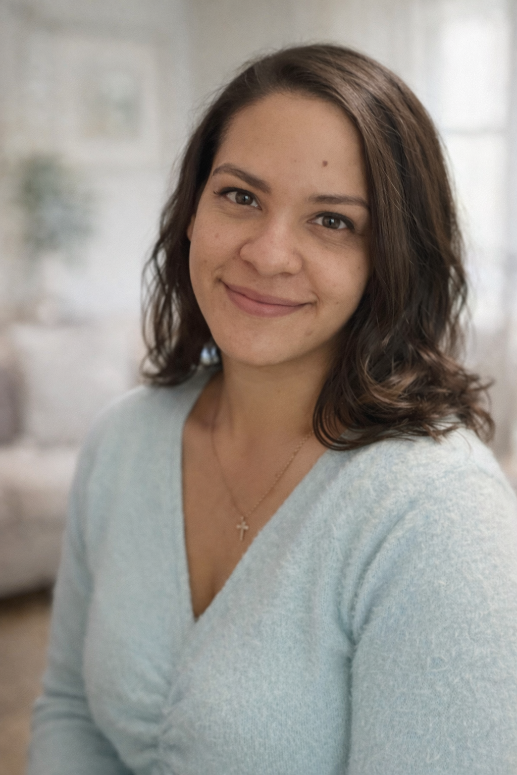 A woman with shoulder-length dark brown hair, wearing a light gray sweater and a cross necklace, smiling at the camera in a softly lit indoor setting.