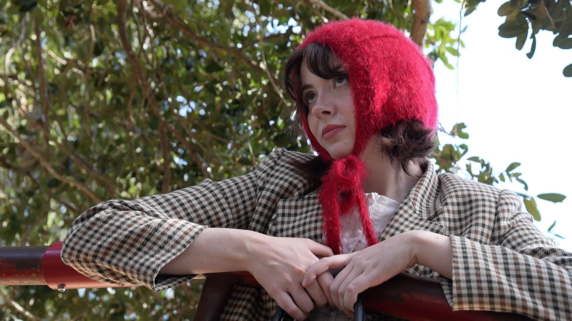 Kadence Roehm, a woman outdoors wearing a red fluffy hat with ear flaps, looking contemplative, with trees and sky in the background.