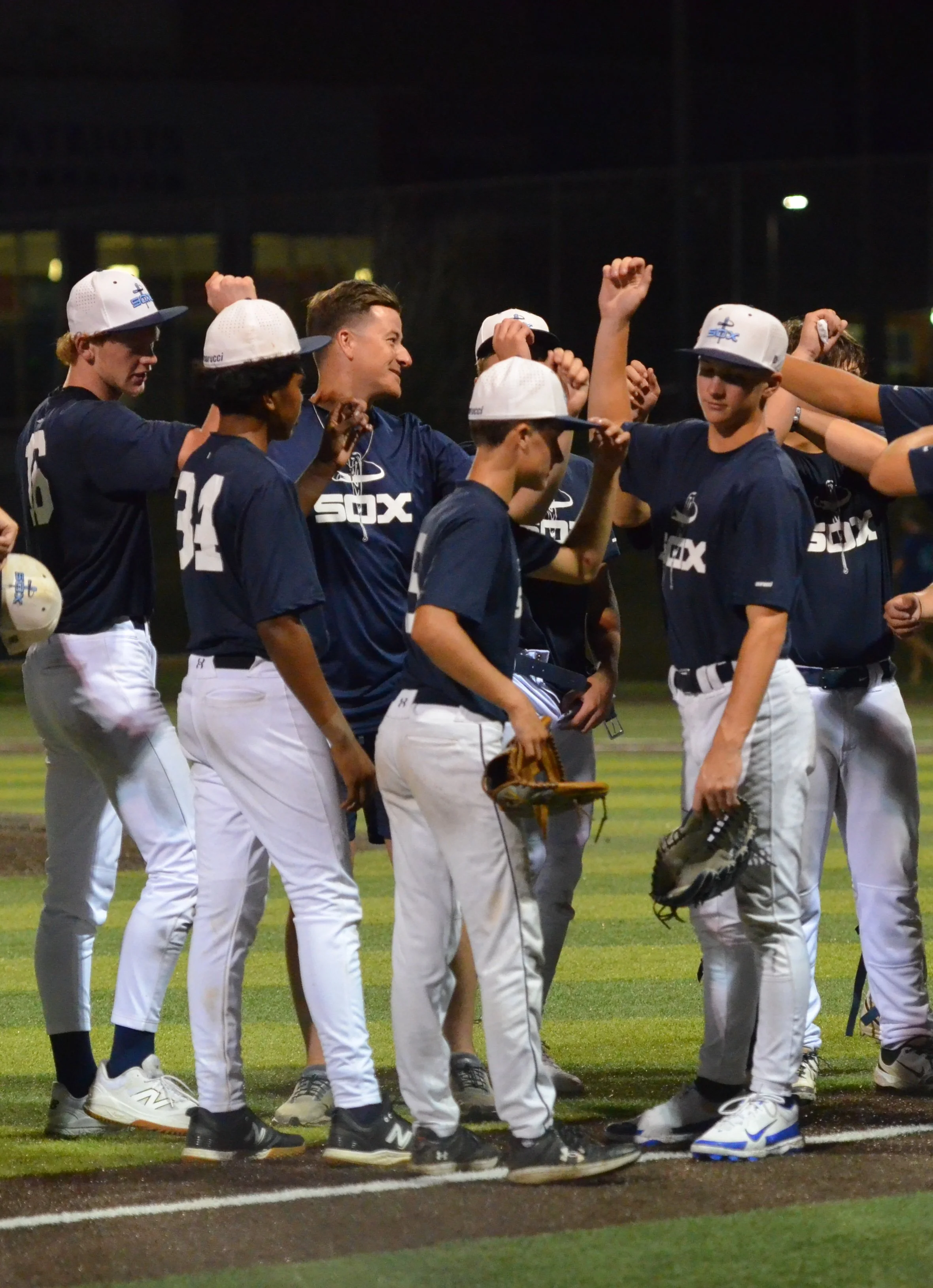 A group of young baseball players, wearing dark blue jerseys with white pants, are standing on a baseball field at night. They are gathered in a circle, some raising their fists, and appear to be celebrating or having a team discussion. One player has a glove in his hand, and they are standing close together.